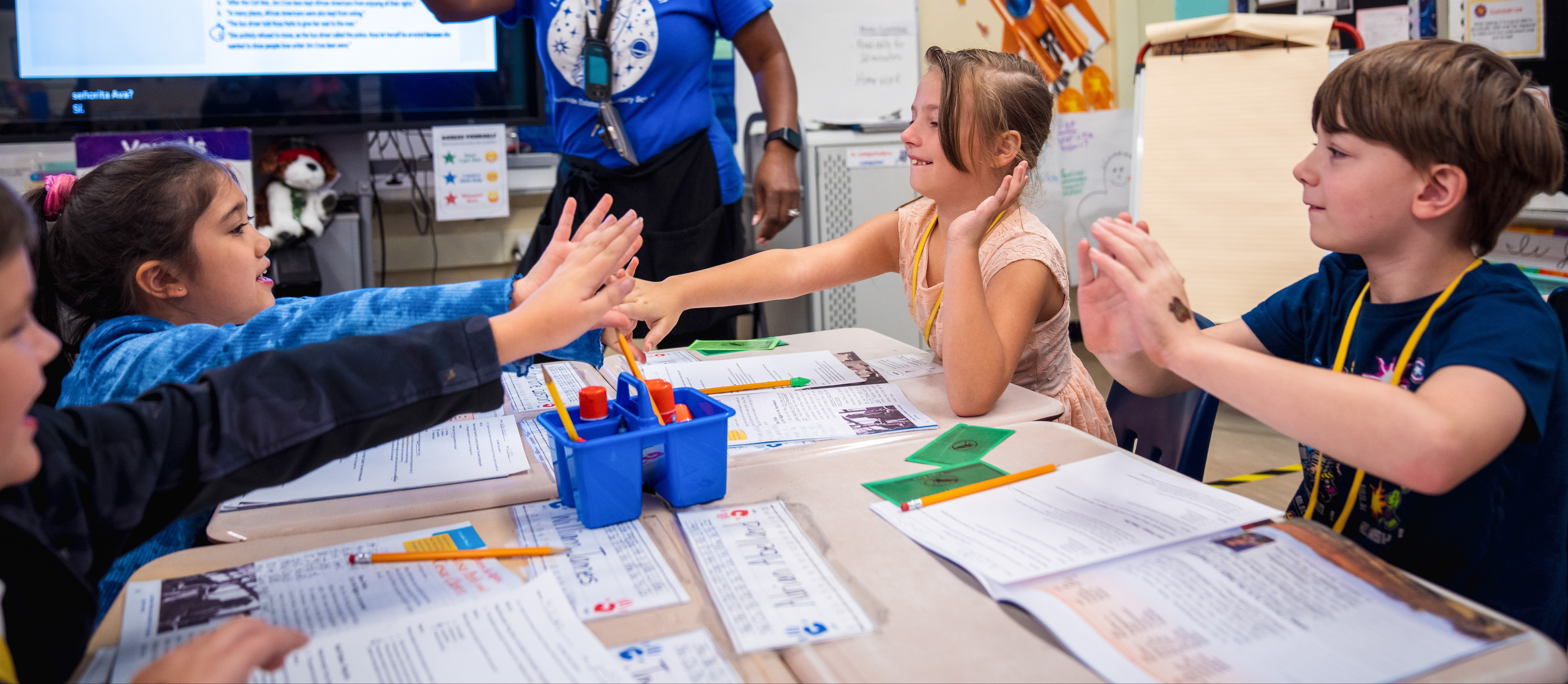 Four children seated around a table, raising hands in class. Papers and pencils on table.