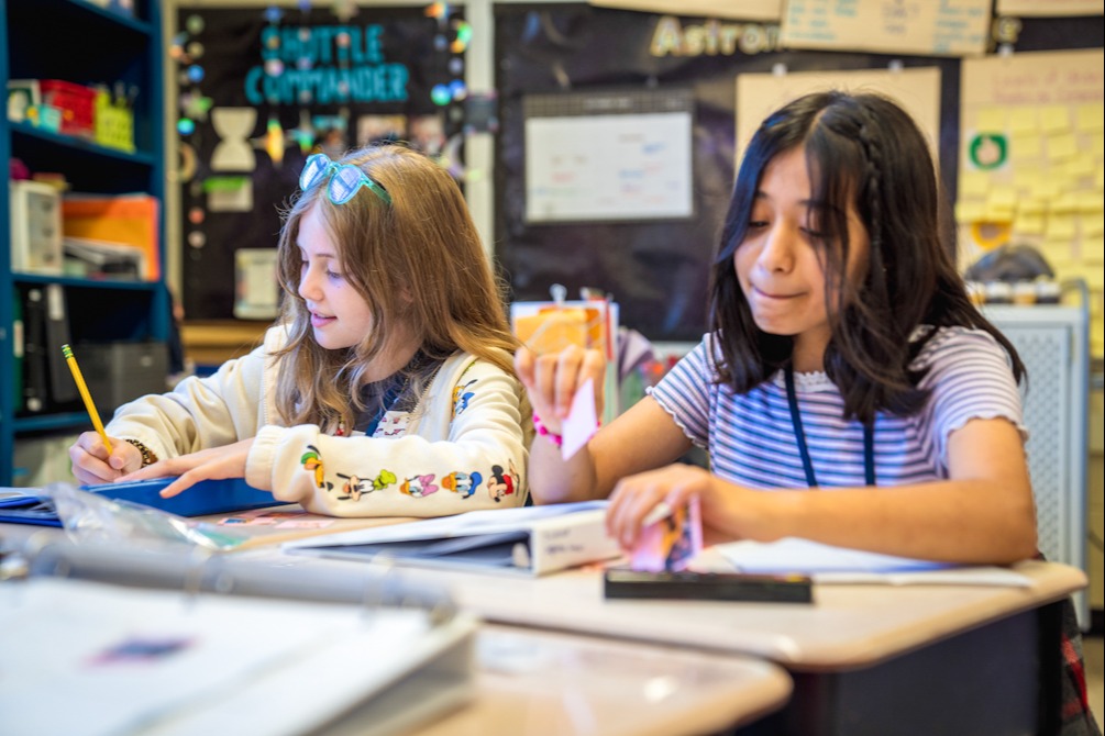 Two children seated in a classroom, one wearing glasses and a headband, the other holding a pencil.