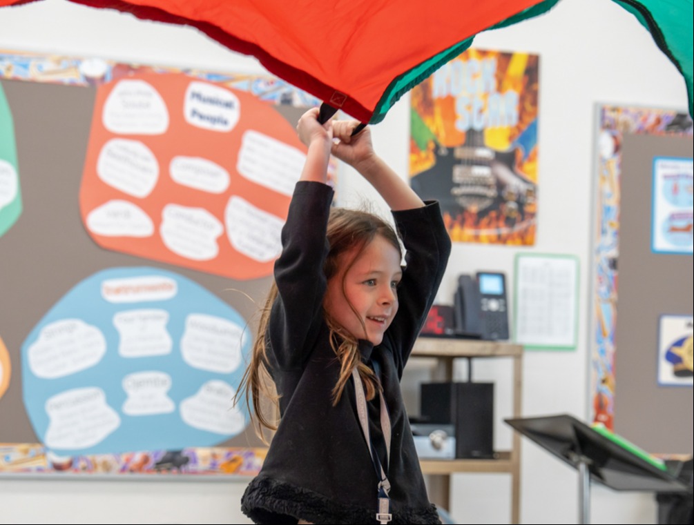A child holds up a colorful parachute indoors. A bulletin board and shelves are behind.