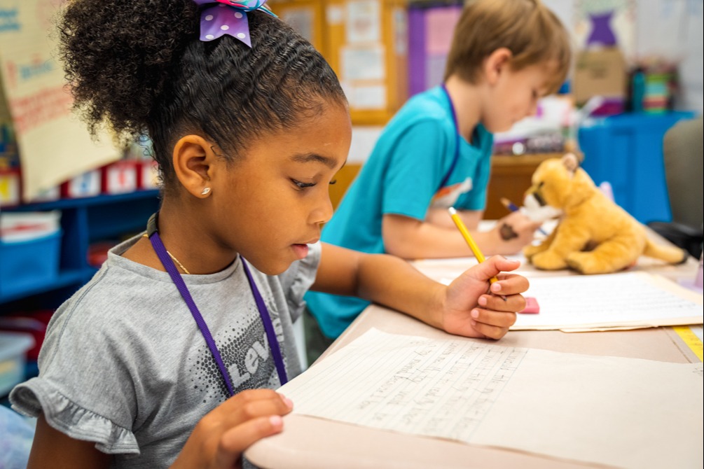 Two students work on papers. A girl writes with a pencil and a boy uses a pen.