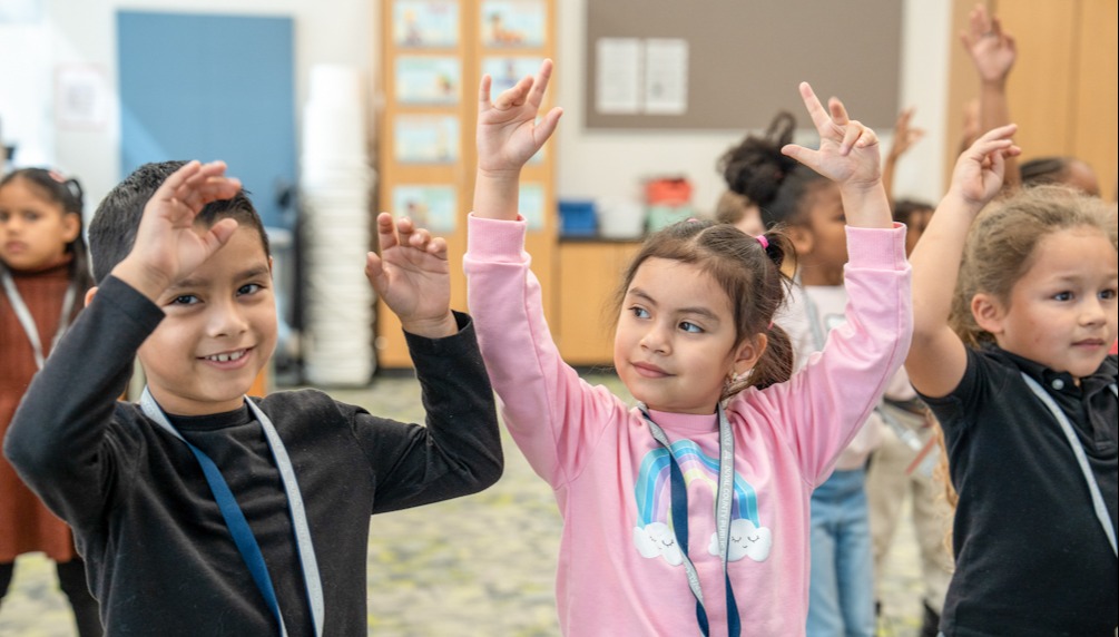 Kids in a classroom raising their hands. One child is wearing a black shirt and another a pink shirt.