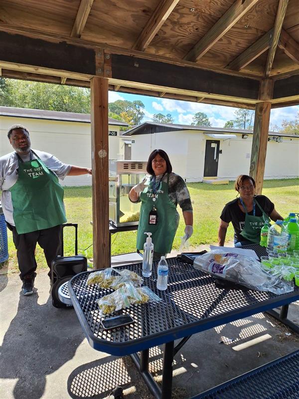 Two people are preparing food at an outdoor table, with a third person standing by, under a covered picnic area. The setting suggests a casual, social gathering.