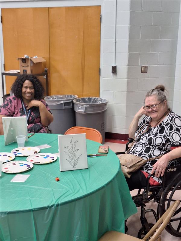 Two women sitting at a table with paintings and cups, in an indoor setting, ready to engage in a creative activity.