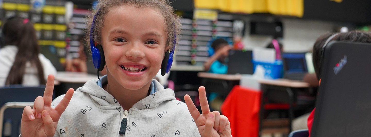 student wearing headphones at a laptop giving two peace signs to the camera