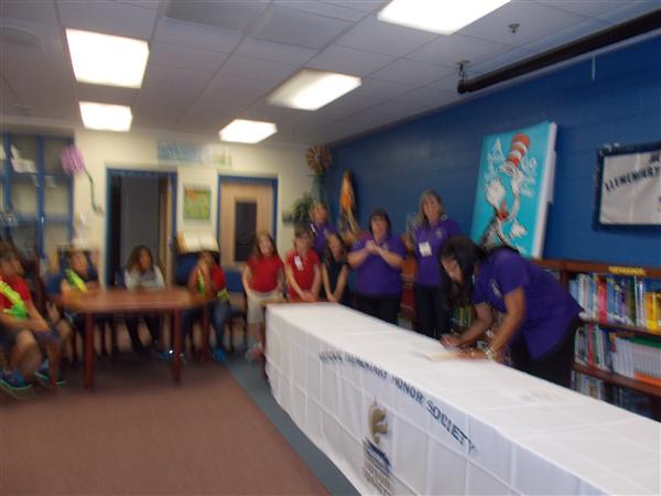 In the image, we can see a group of people gathered in a room with a blue carpet. Some individuals are standing around tables, while others are seated. The focal point appears to be a long table covered with boxes and papers. One person is signing a document atop these materials. A sign in the background displays text which is not entirely legible; however, it seems to be related to some sort of school or educational institution.
