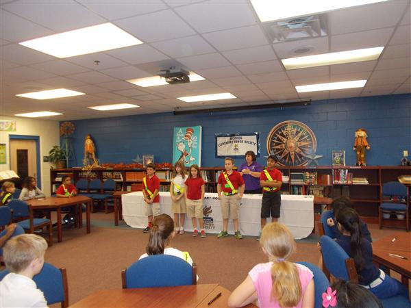 This is a classroom scene where students are standing in front of desks, and there's a presentation happening with a woman speaking to them.