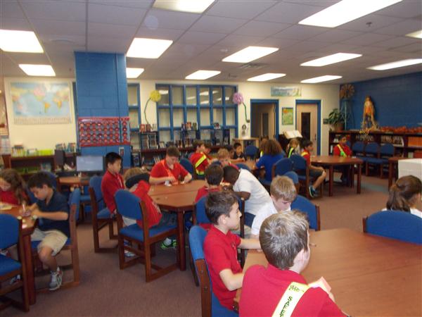 A group of students, both seated and standing, working on a project in a classroom setting with a teacher present.