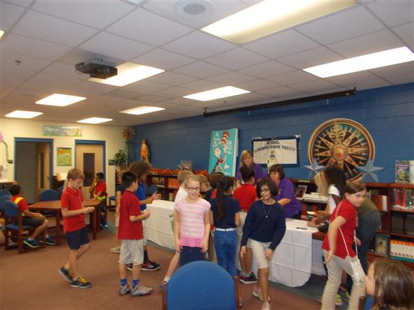 Group of children gathered in a library, engaged in various activities on laptops and tablets.
