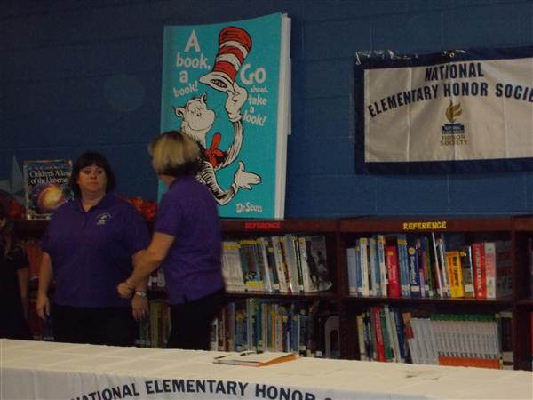 Two individuals in a room at an event, with one person standing behind a table adorned with books and a poster.