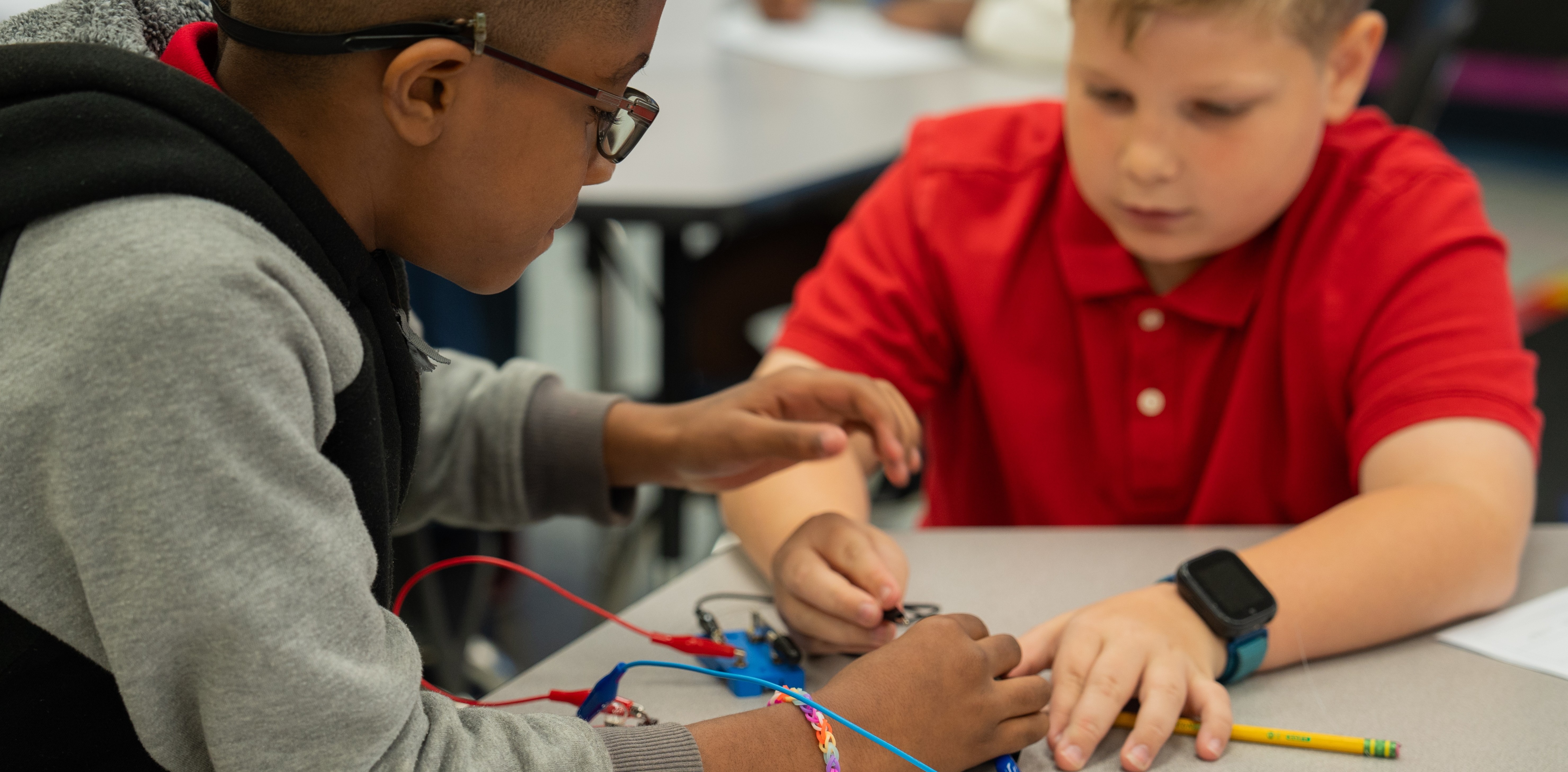 Two boys with glasses, one in a red shirt, the other in a gray, work on an electronic project.