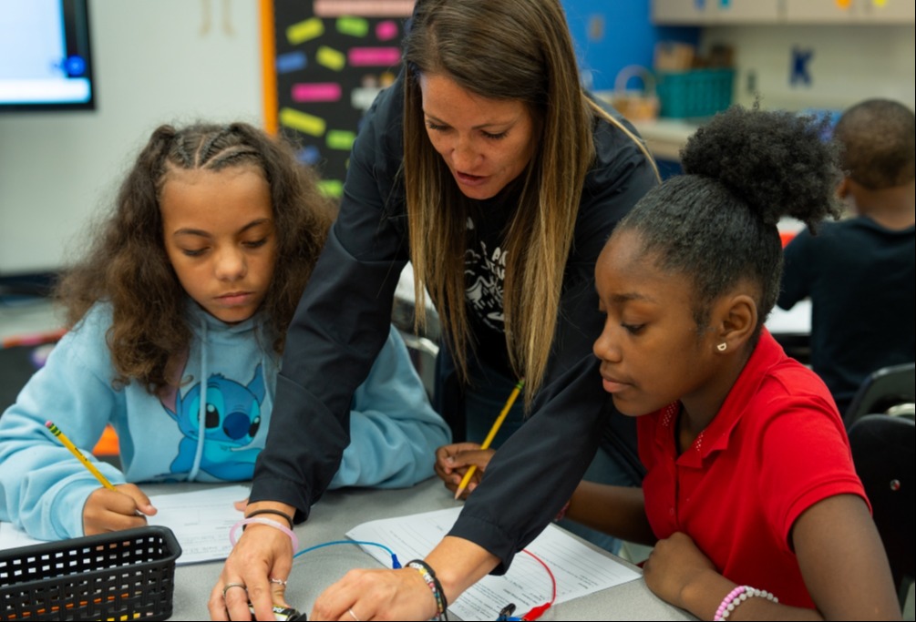 A teacher assists two students with their work at a desk in a classroom.