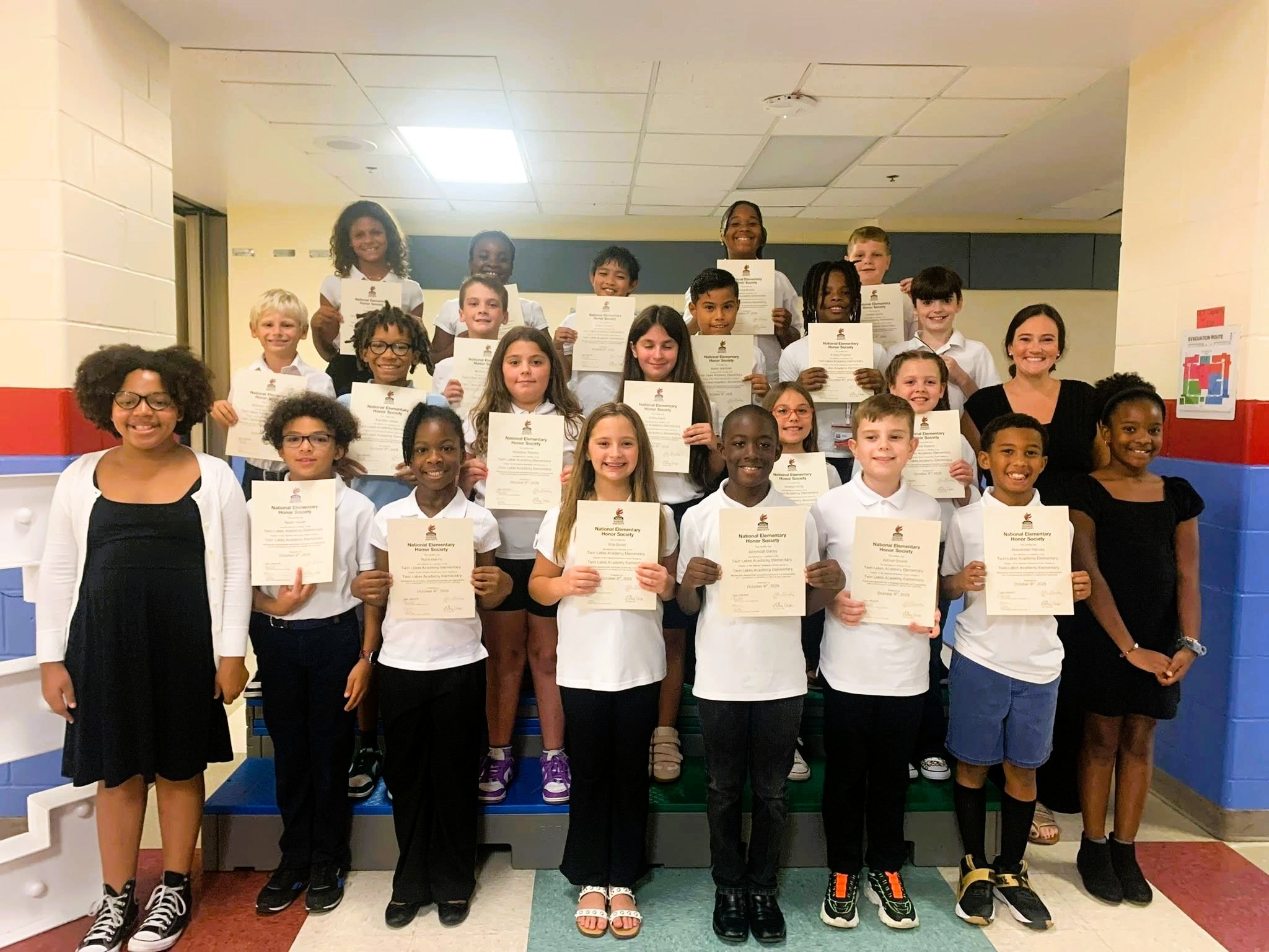 A group of young students in a hallway hold certificates while standing in front of a teacher.