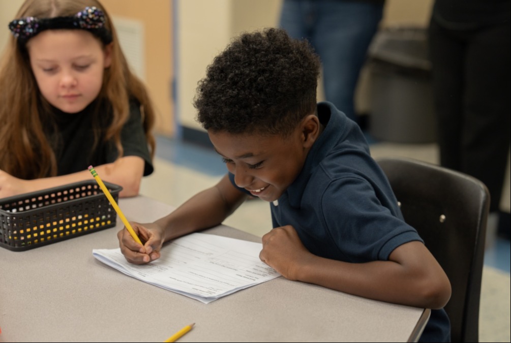 Two children seated, one writing with a pencil, another observing. A desk holds paper and pencils.