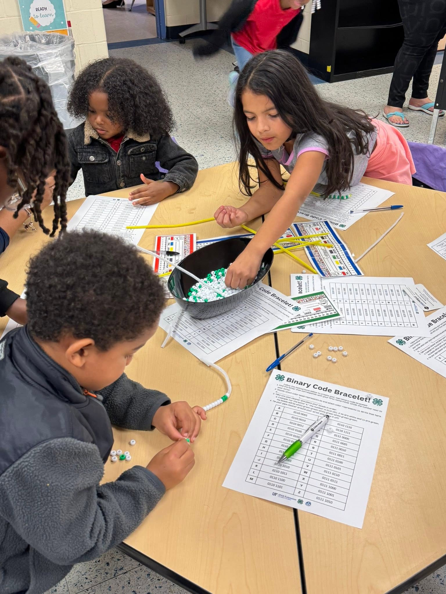 Four children at a table, working on a project with paper and beads. One girl is reaching into a bowl.