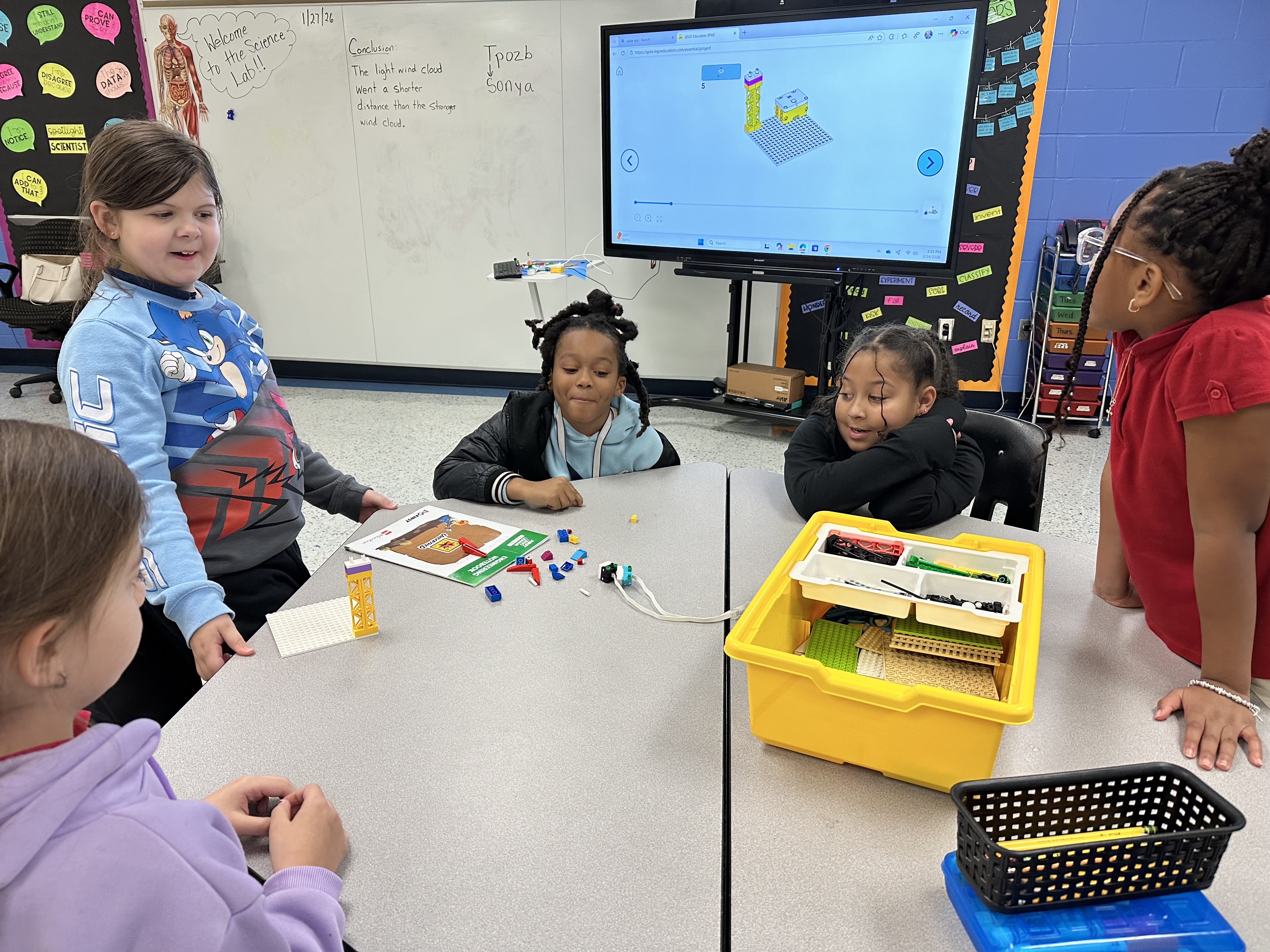 Children in a classroom gather around a table with books, electronics, and a monitor on a stand.
