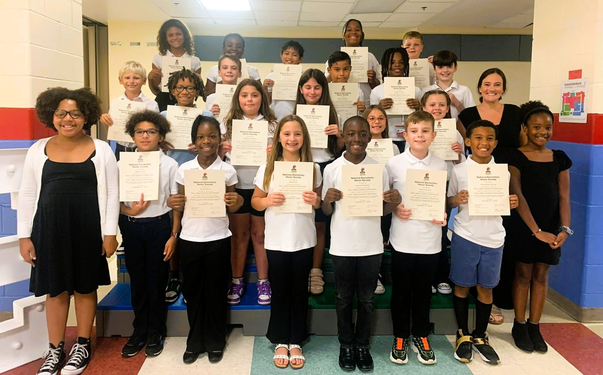 A group of children in white shirts and various pants, some holding certificates, stand in a hallway.