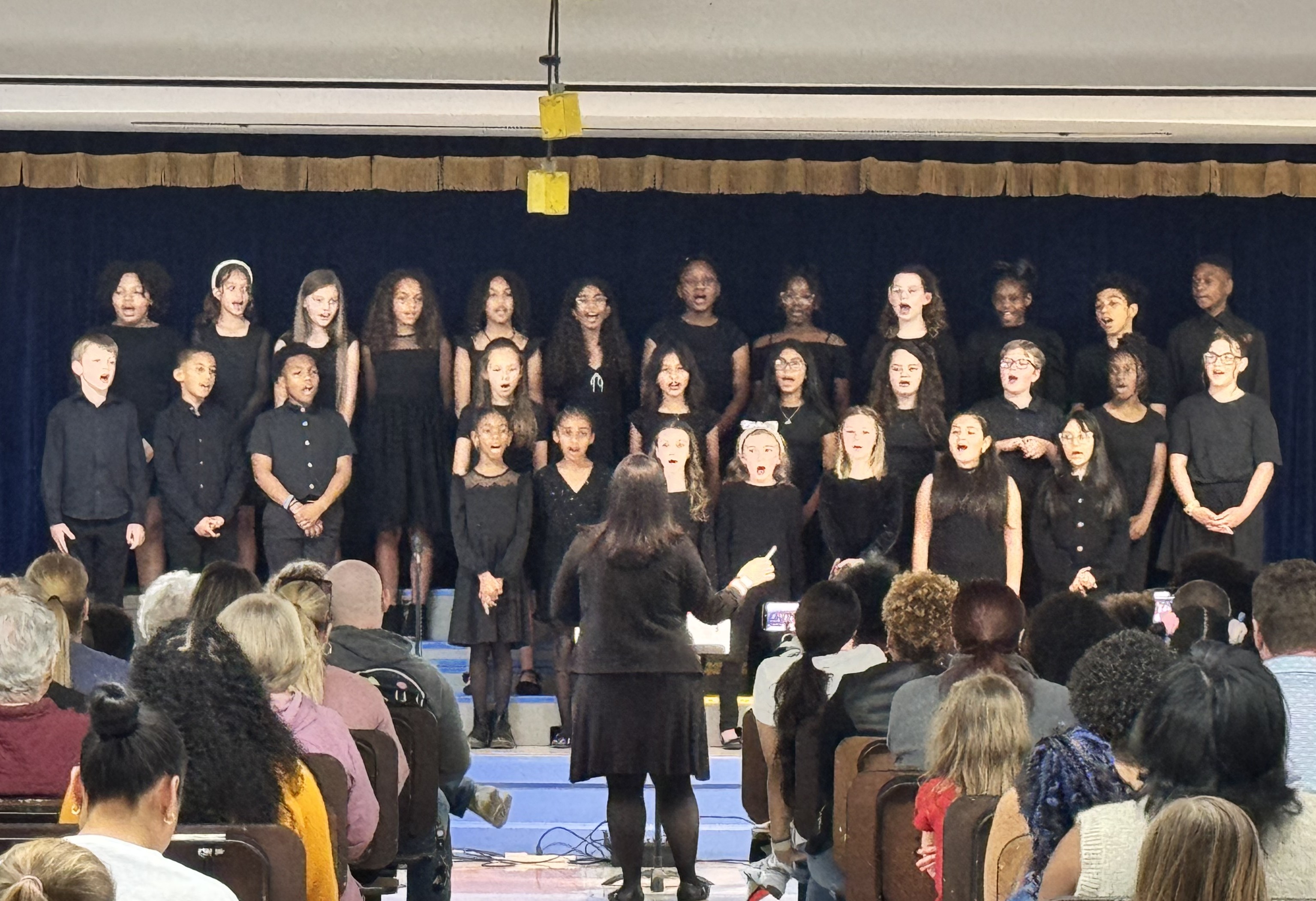 Choir members in black attire perform on stage with a blue backdrop, while a conductor directs them. Audience members sit in rows, attentively watching.