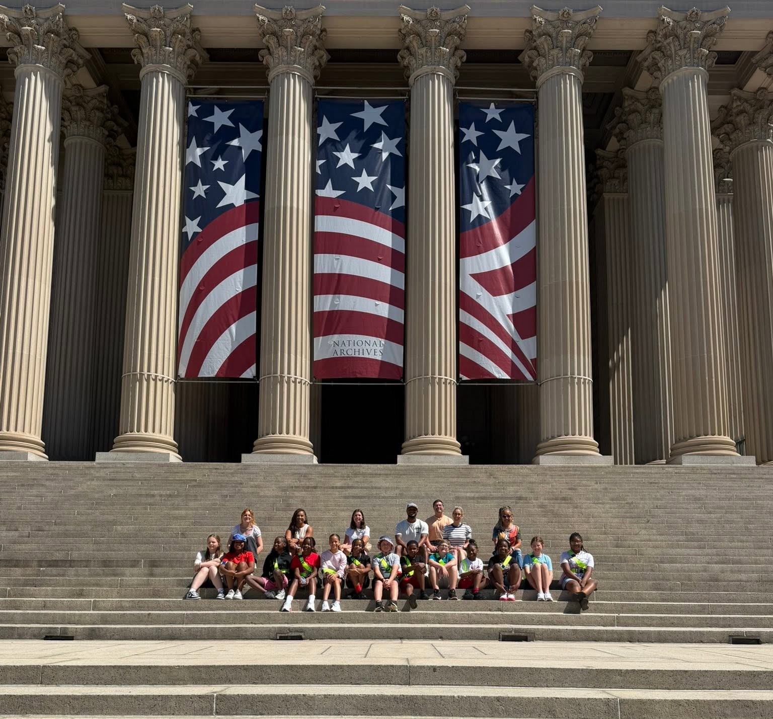 A group of people in uniforms poses on the steps of a building with two large American flags.