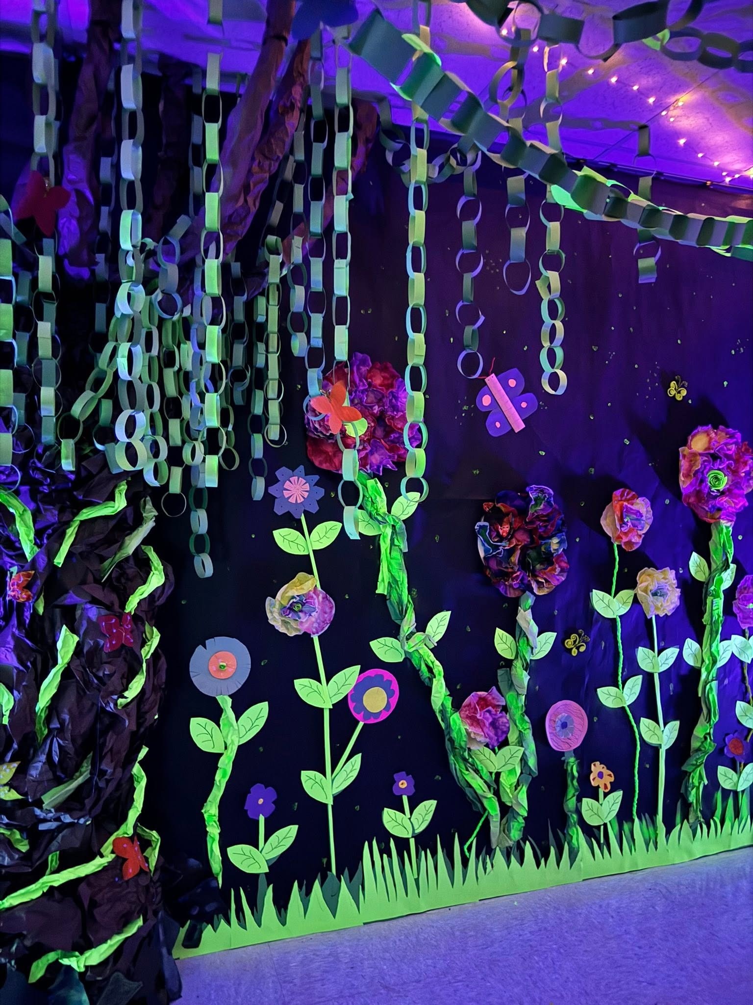 A colorful display with flowers, vines, and glowing elements on a dark background. Chains hang from above.