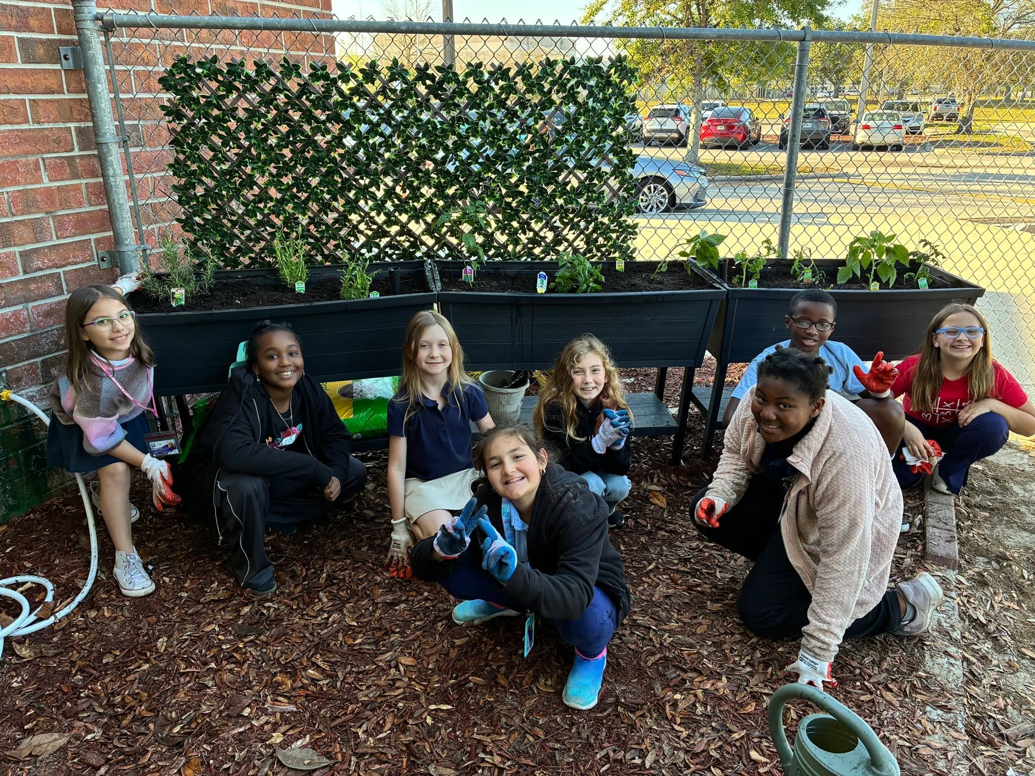 Students and adults crouch in front of a raised planter in a garden with a chain-link fence.