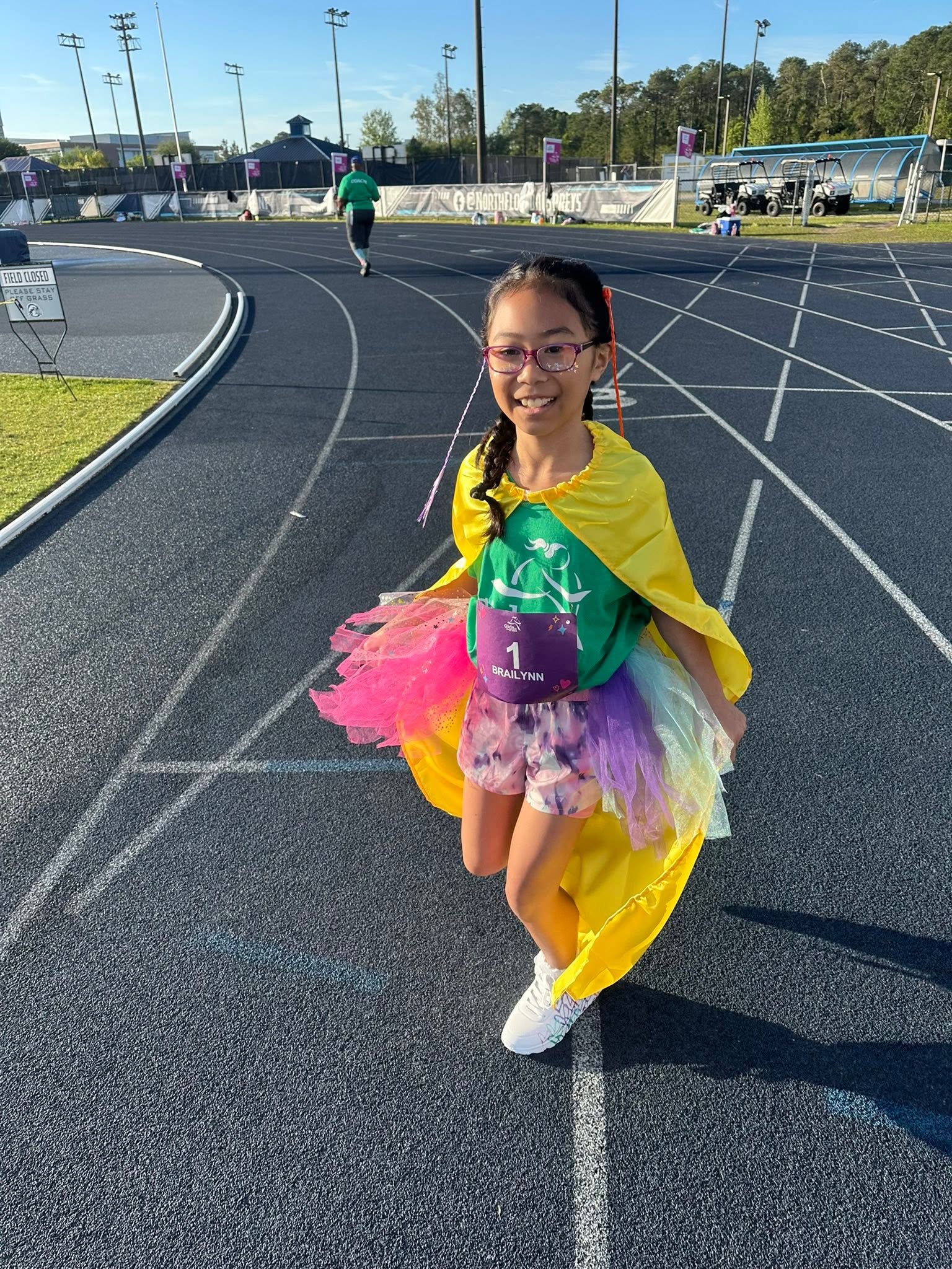 A young girl in a superhero costume runs on a track. She wears glasses and holds a cape.
