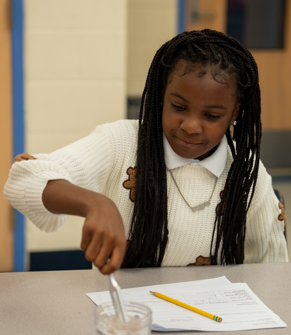 A young girl is seated at a desk, working with a pencil and a jar, wearing a white sweater.