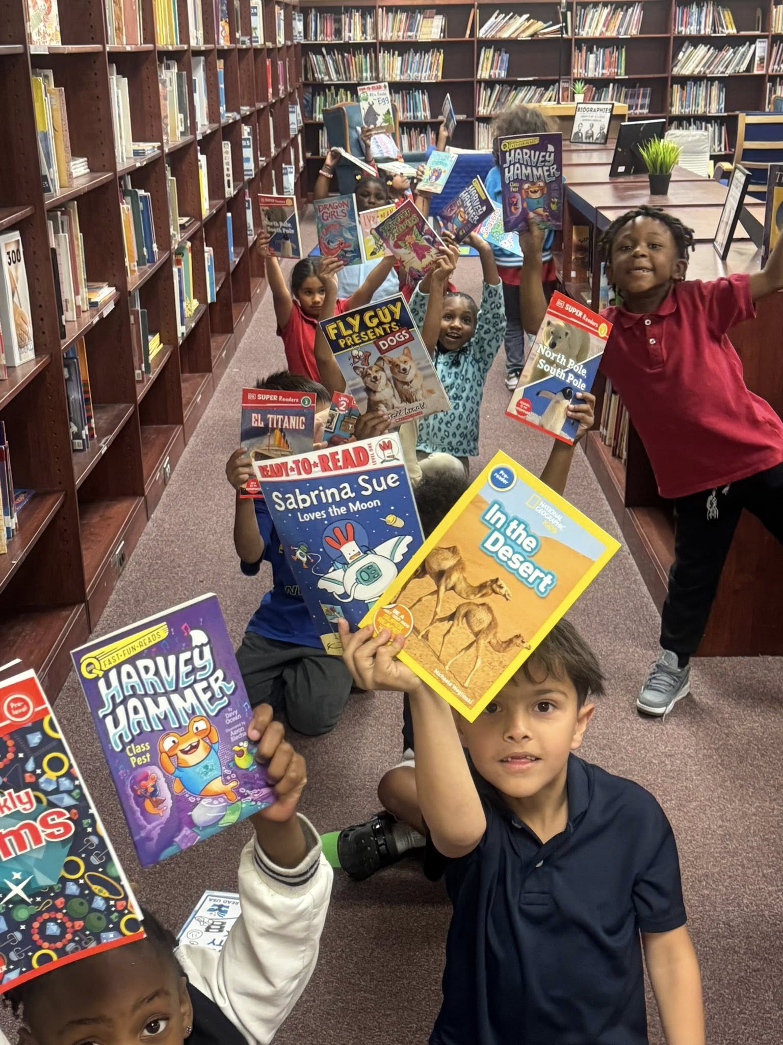 Kids holding up books in a library aisle, with shelves of books in the background.