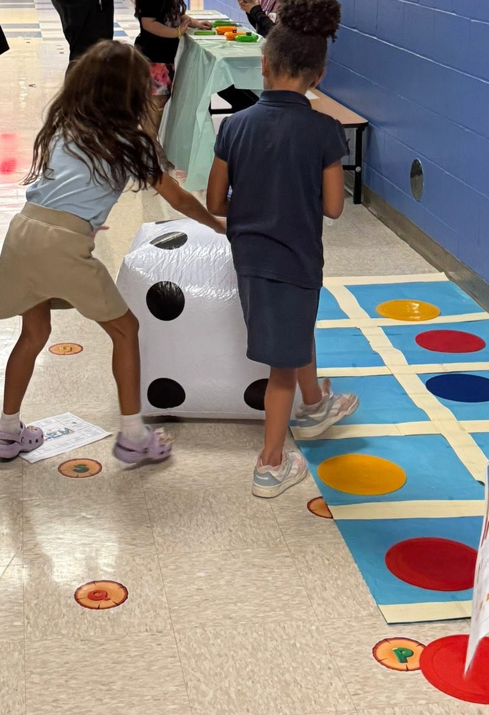 A hallway with children playing a life-size math game. Colorful dice and circles on the floor. Some individuals stand behind tables.