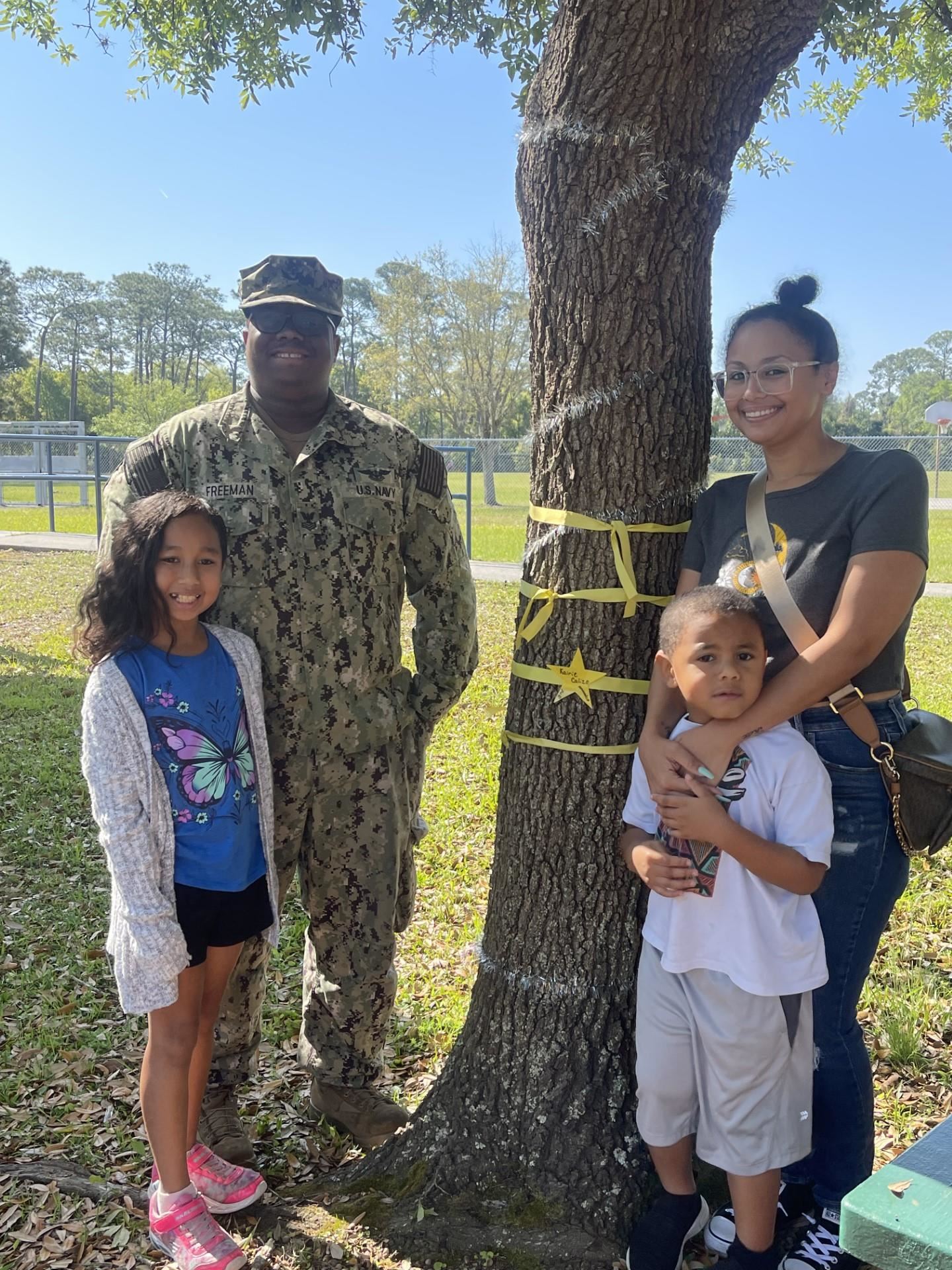 Military personnel, family members and civilians posing near a tree with ribbons.