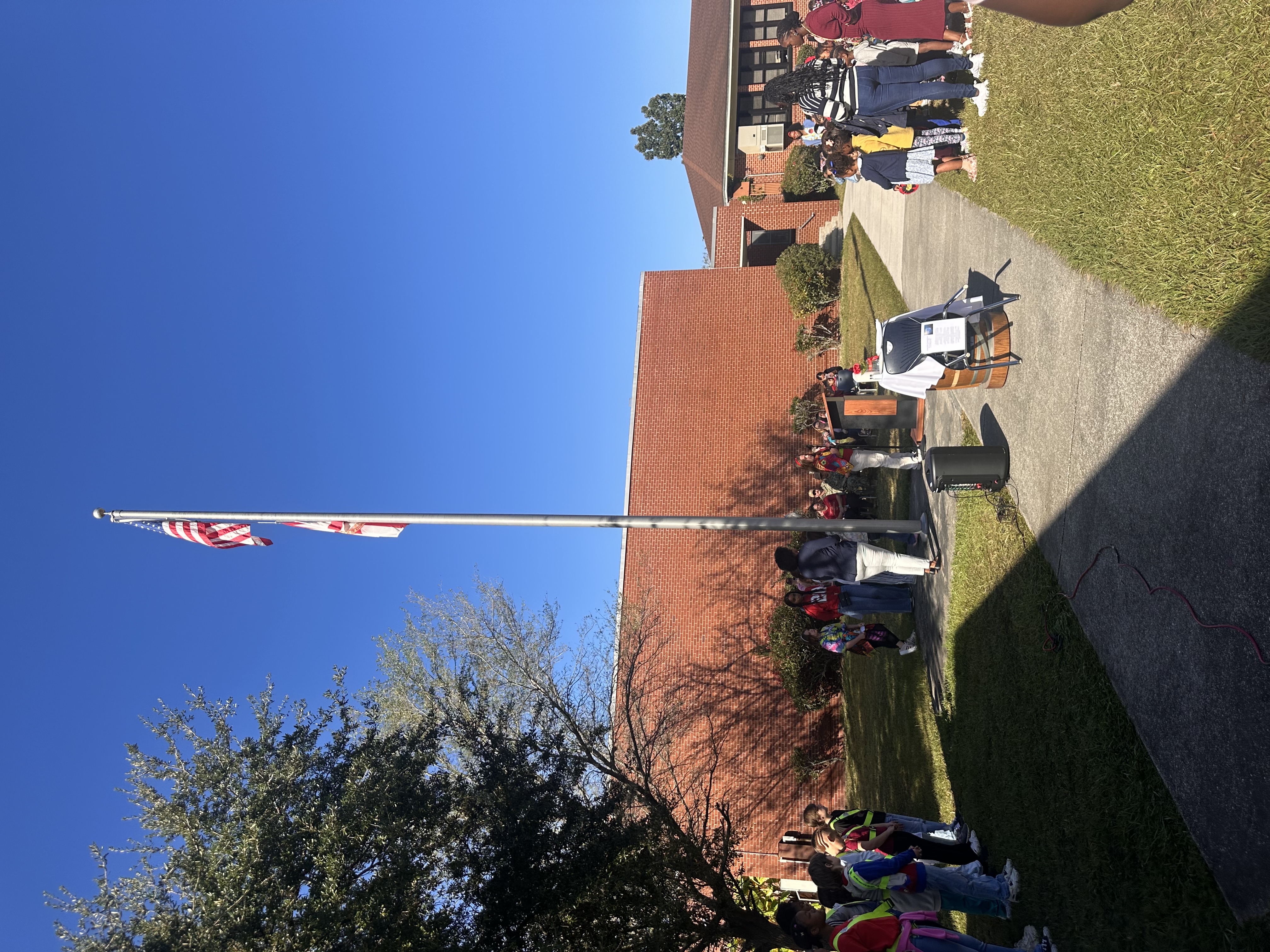 People stand in a line on a sidewalk, near a flagpole, in front of a red building.