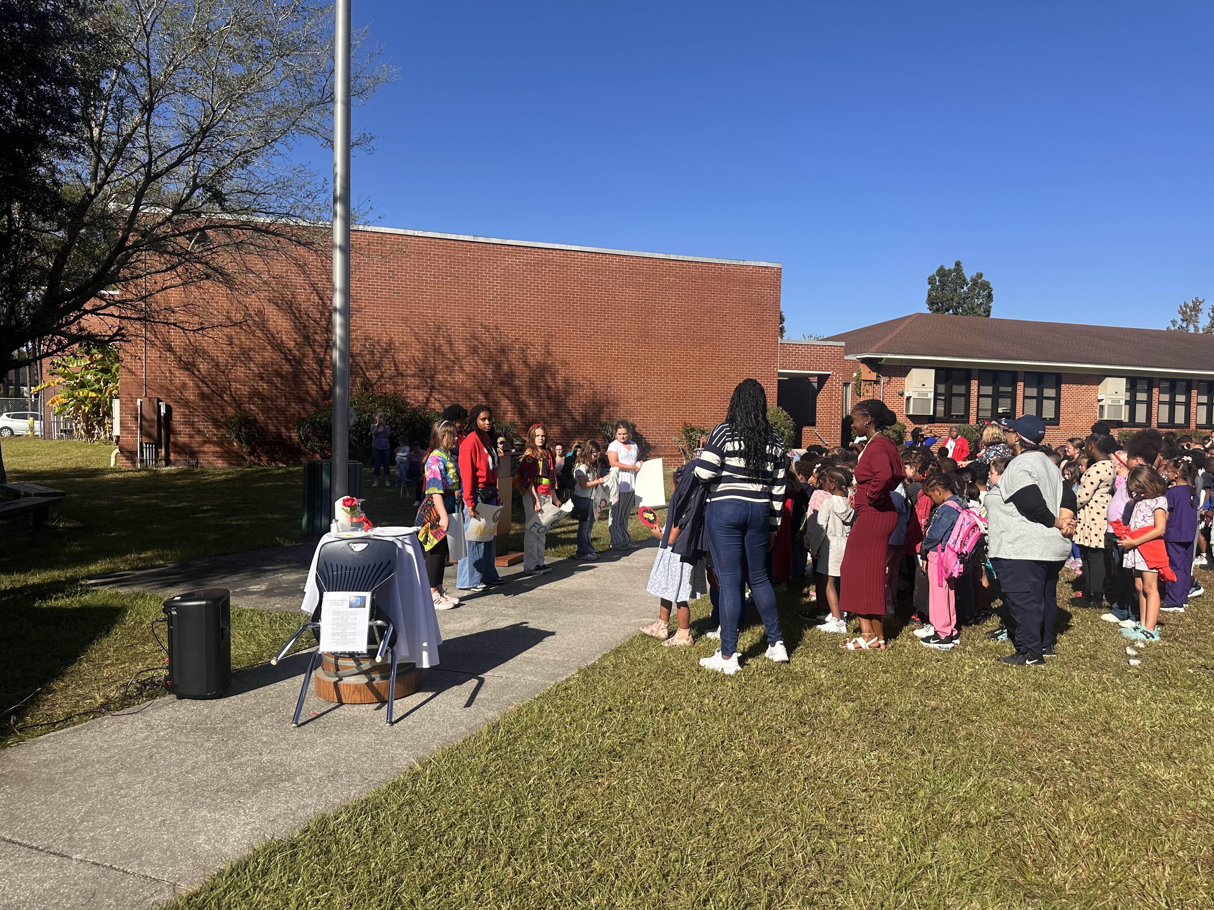 Group of people outside in a line, facing a building with a flagpole, standing on a grassy lawn.