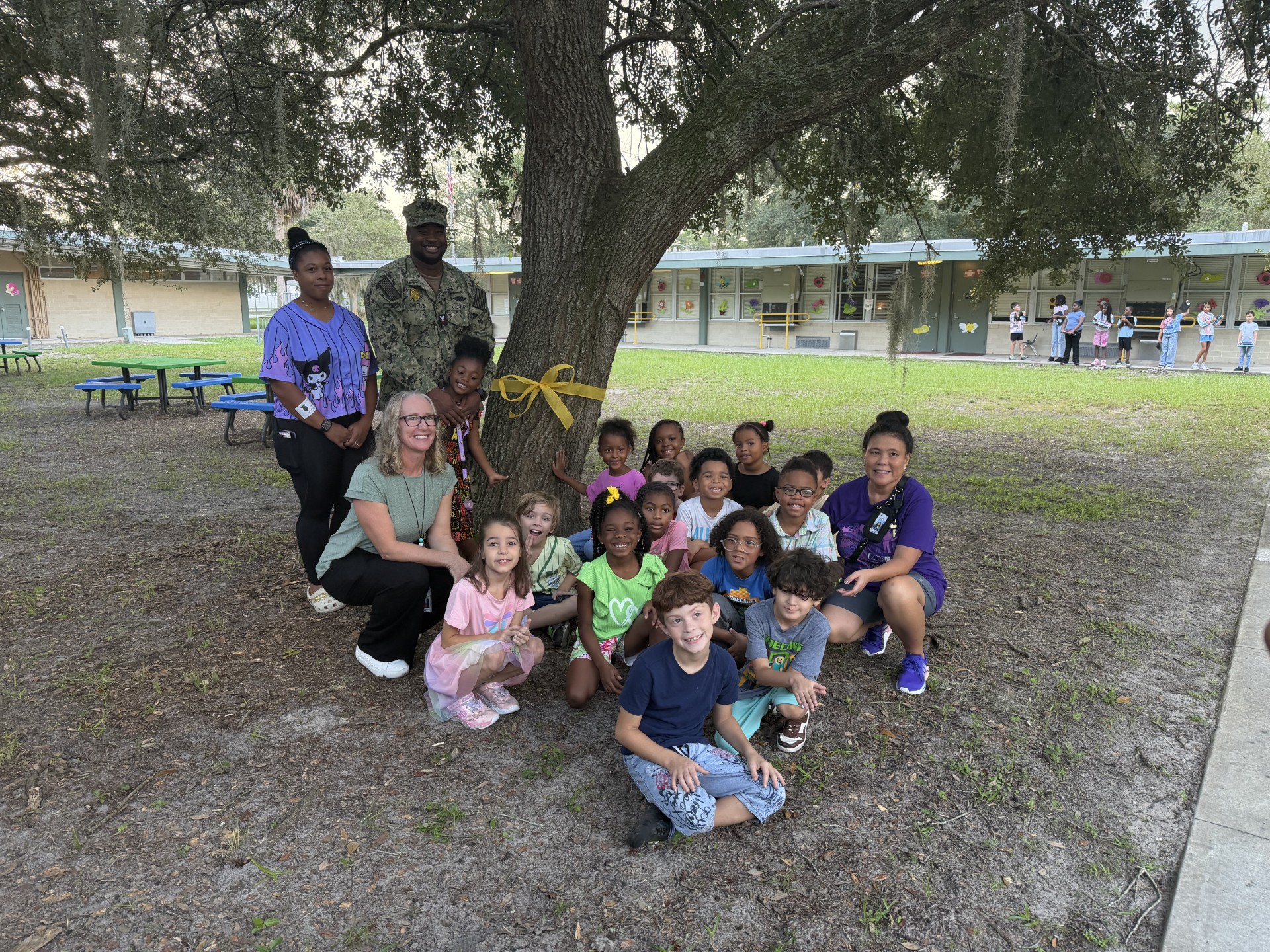 A group of children and adults pose under a tree with a yellow ribbon in a park.