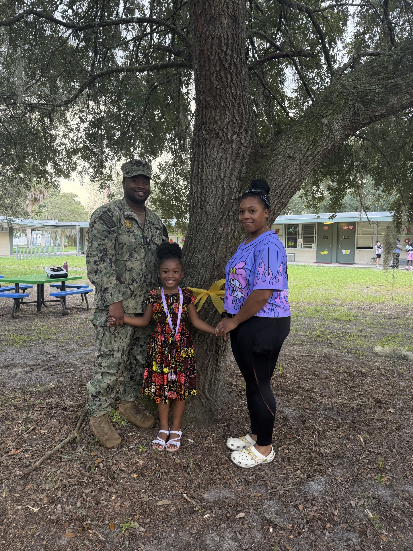 Man in uniform, woman, and child standing under tree, posing for photo with ribbon tied around tree.