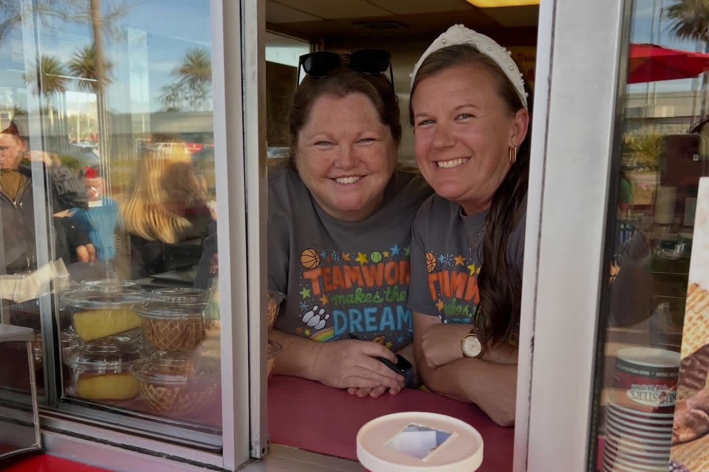 Two women smile, one with sunglasses on her head, sit at a food stand counter with ice cream and plates.
