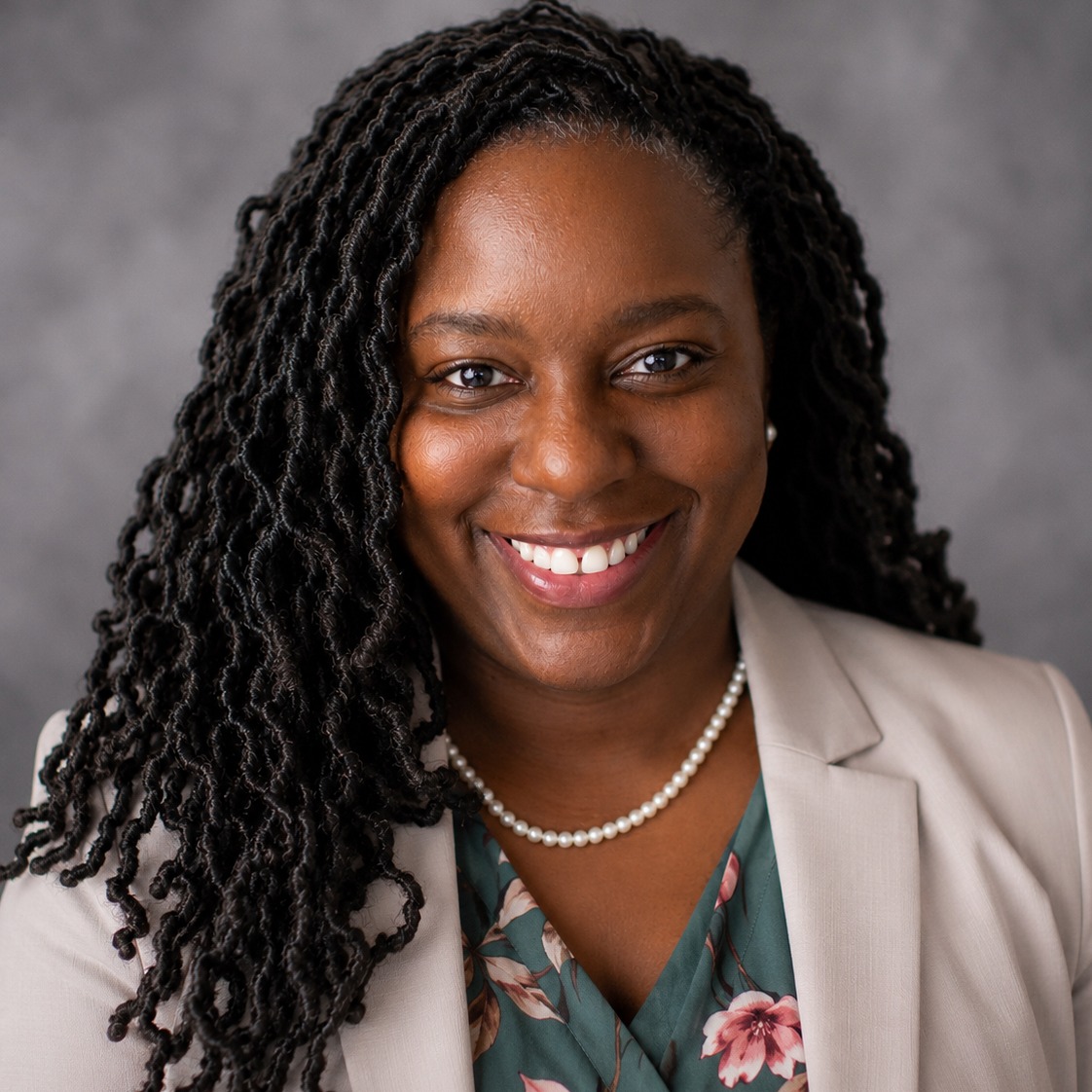 Woman with curly hair, smiling. Wearing a white blazer over a green floral shirt and a pearl necklace. Background is gray.