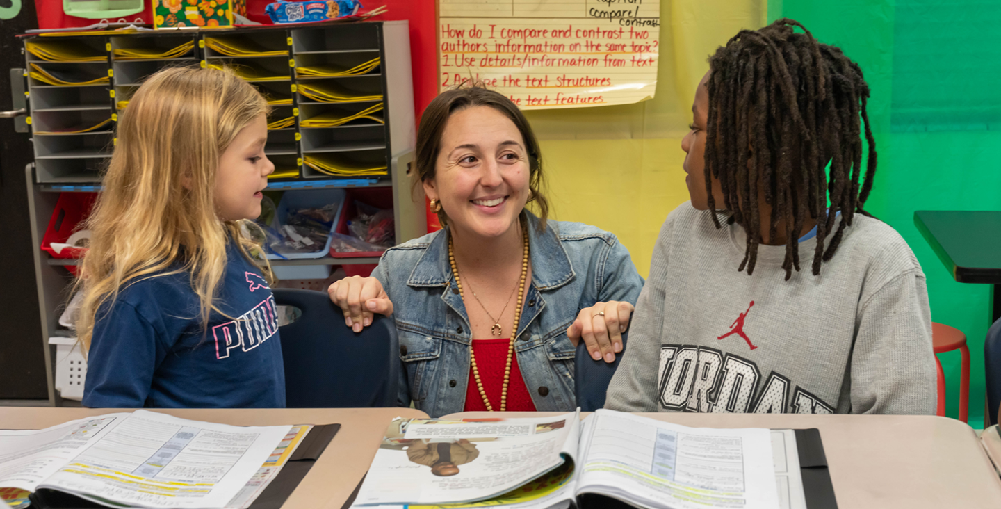 Teacher kneeling to talk to two students at a desk