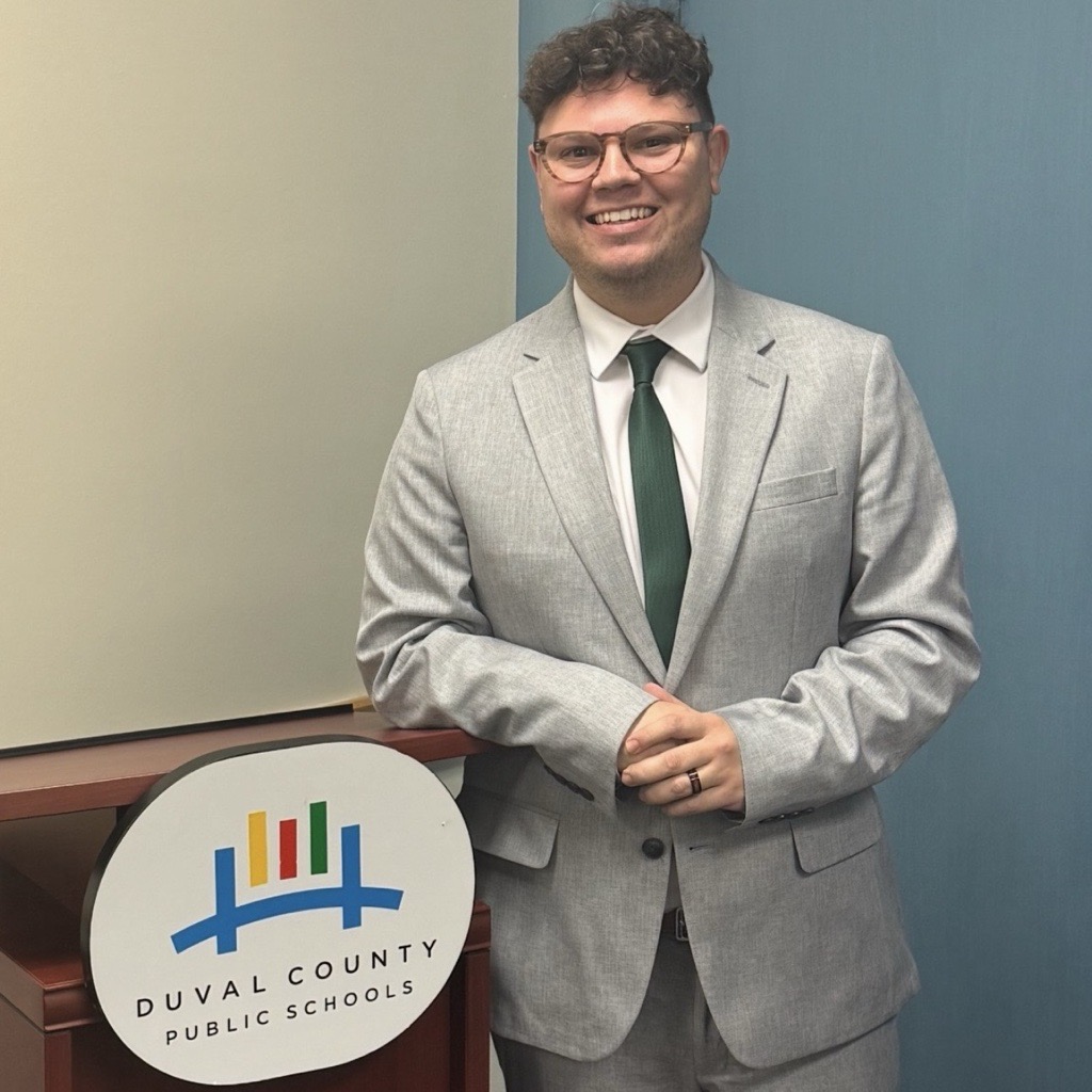 A man in a gray suit and tie smiles, standing near a podium with a colorful logo.