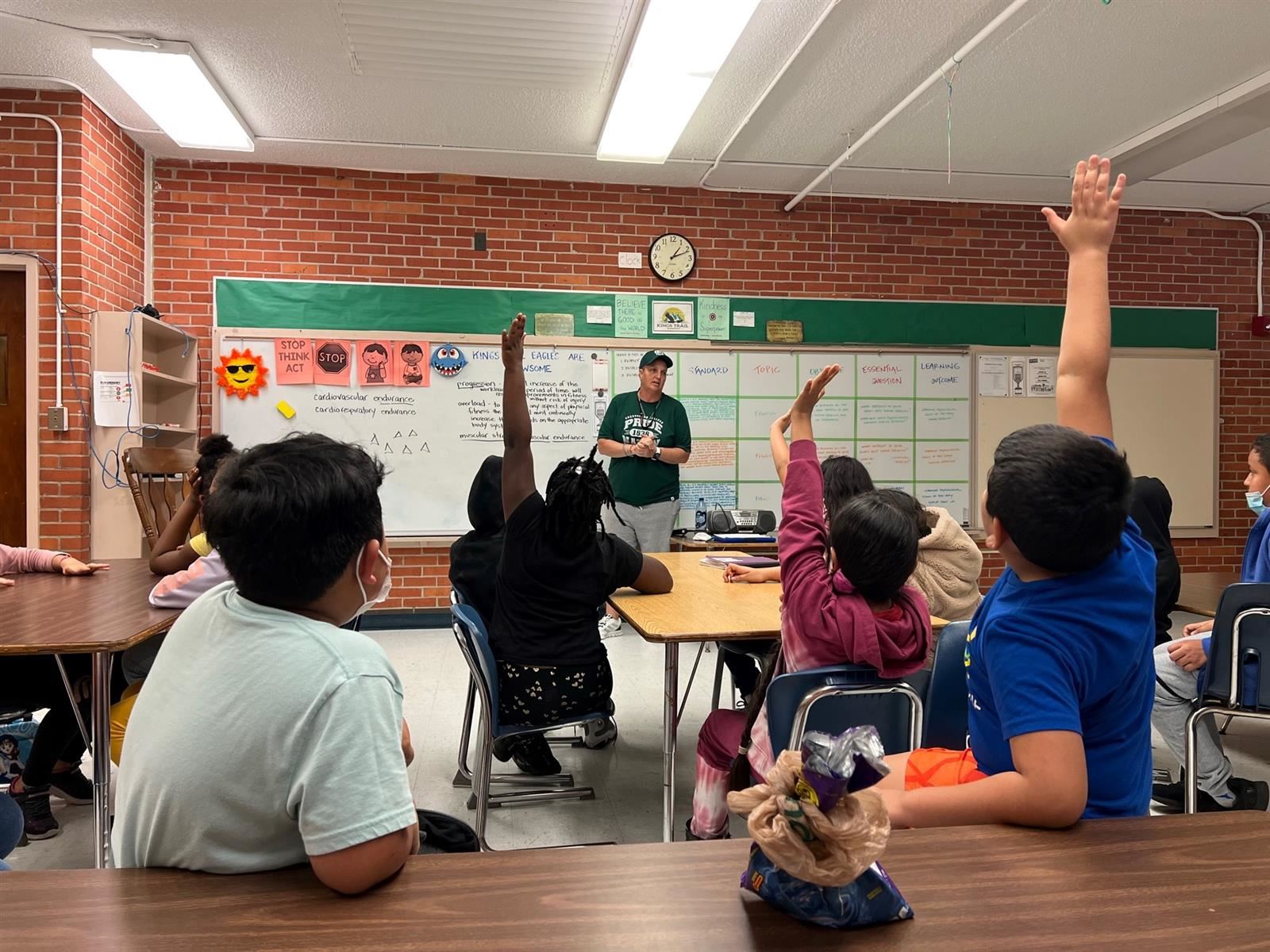 This is a photo of a classroom setting with a teacher standing at the front and students seated in rows, engaged in an educational activity.