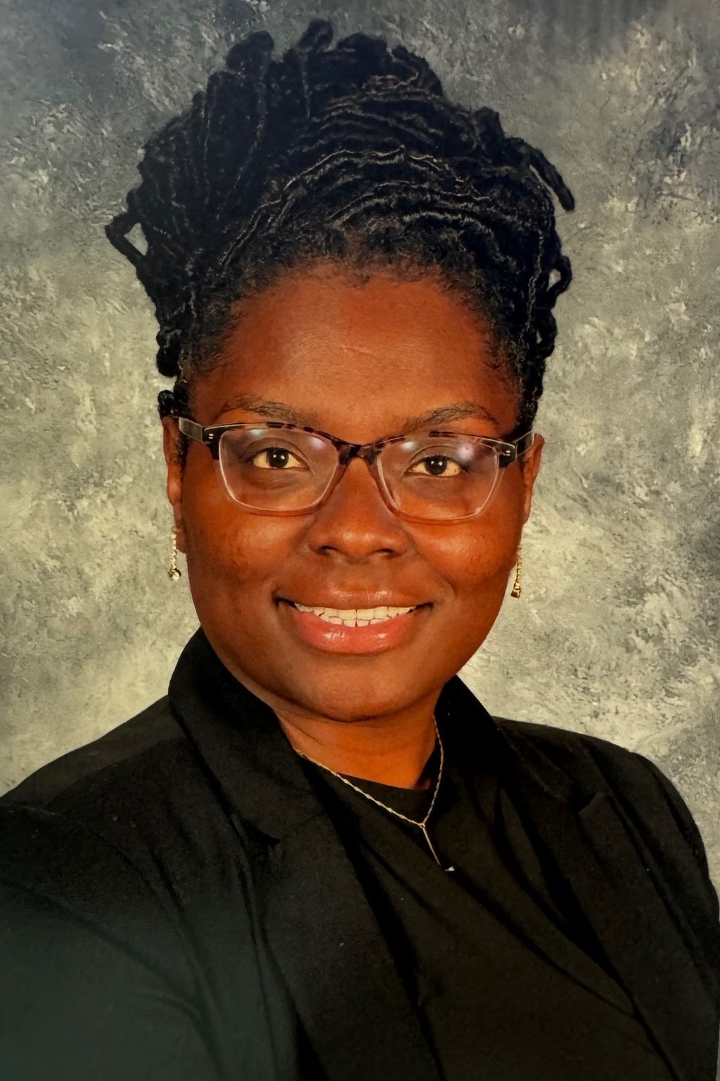 A woman with glasses and a black shirt, smiling at the camera. Her hair is styled in braids.