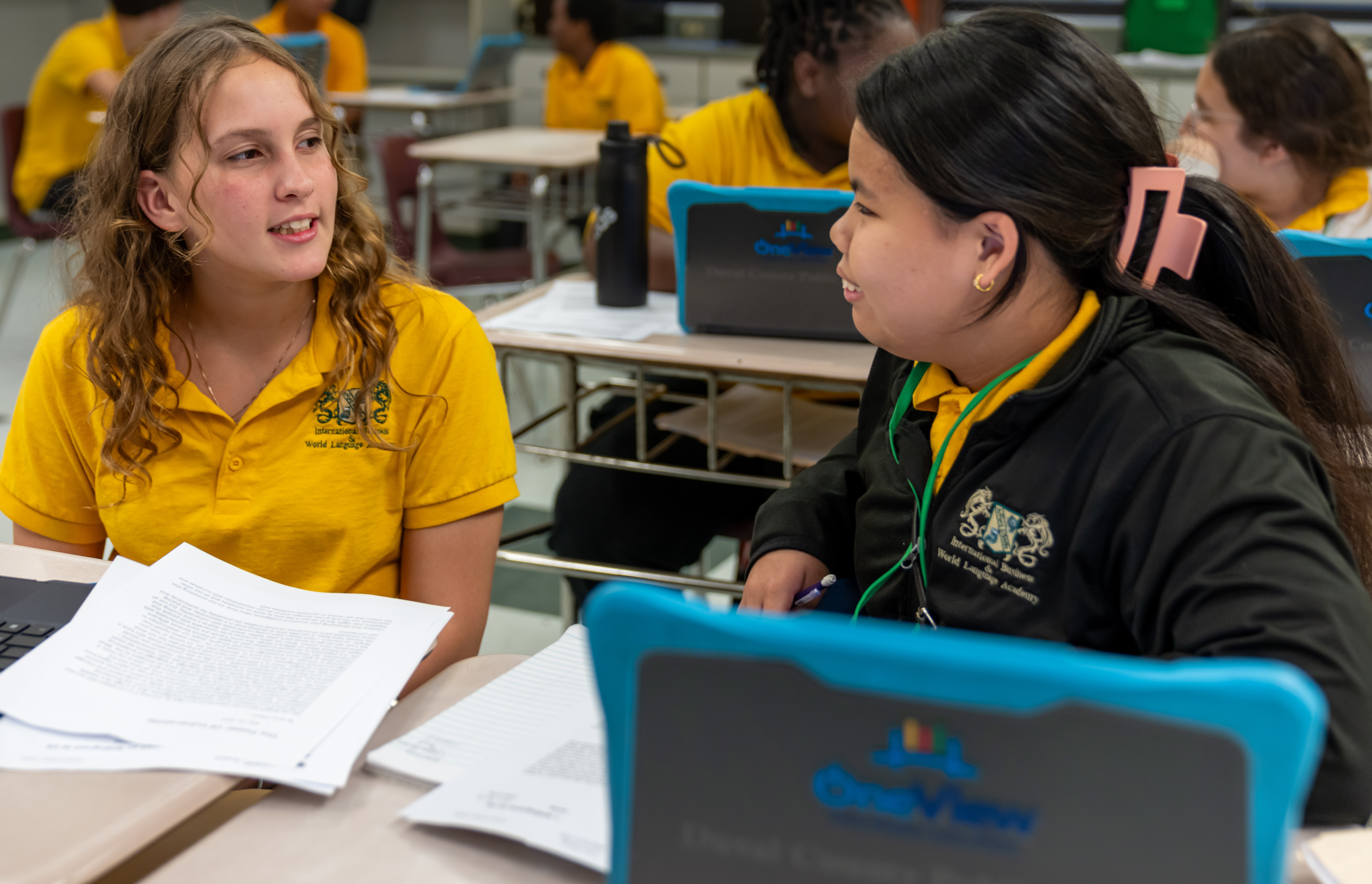 Two girls in a classroom with laptops, one wearing a yellow shirt and the other in black.