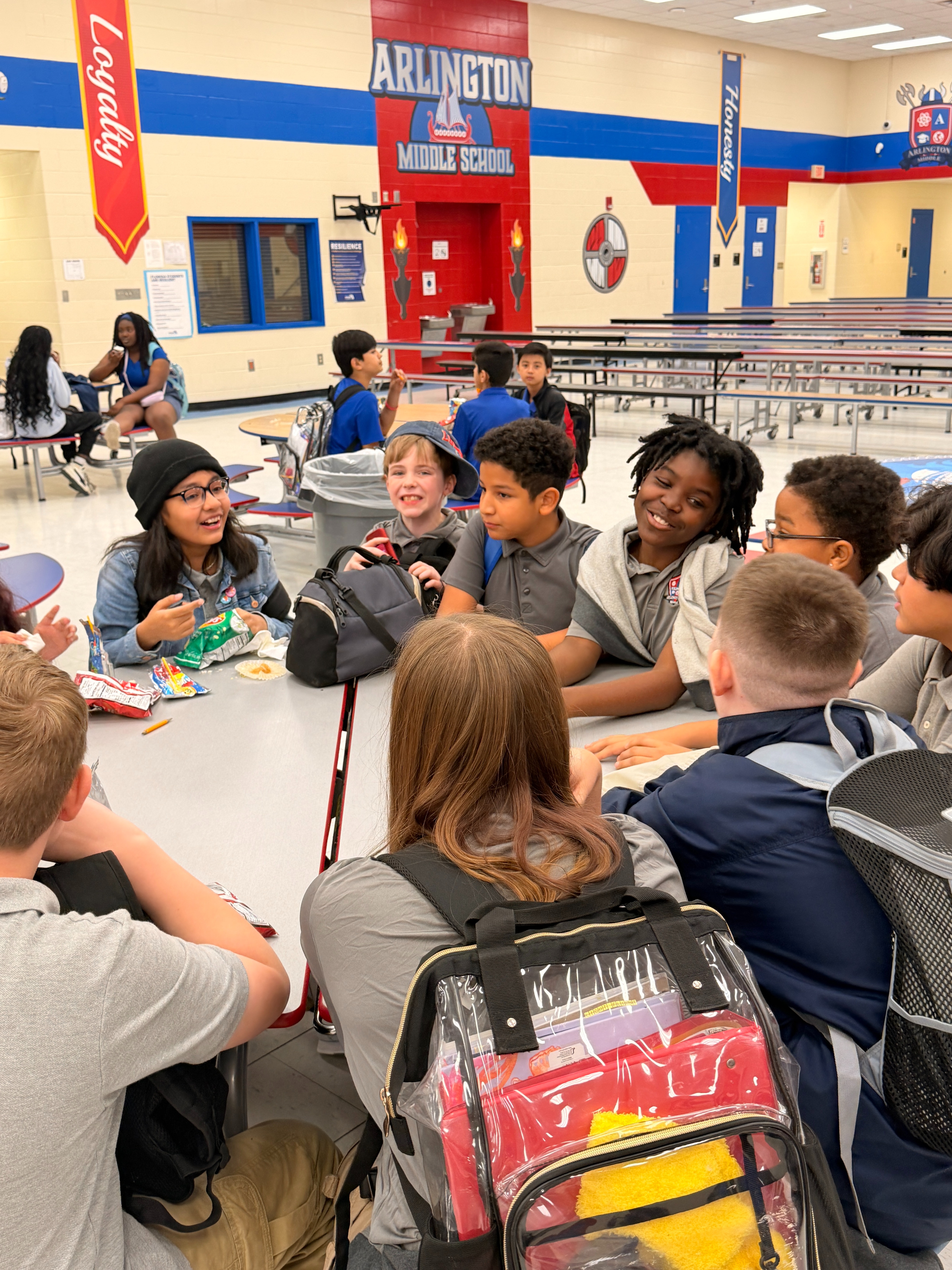A school cafeteria with students sitting at a long table. Some wear backpacks, and others have bags on their laps.