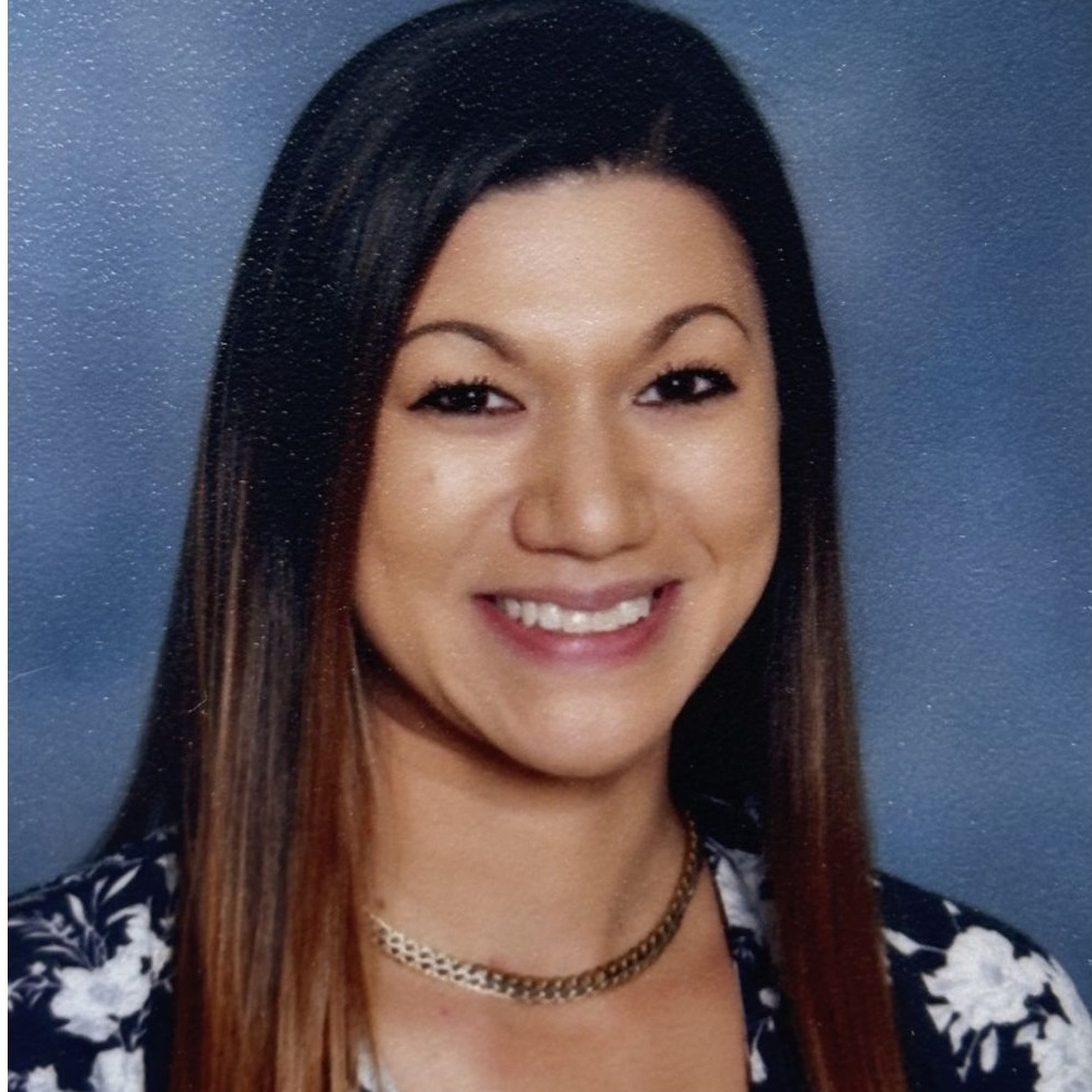 A smiling woman with long, straight dark hair, wearing a black and white floral blouse and a necklace, poses against a blue background.
