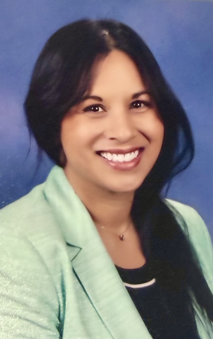 A woman with dark hair smiles for the camera. She wears a black top and a mint-green blazer.