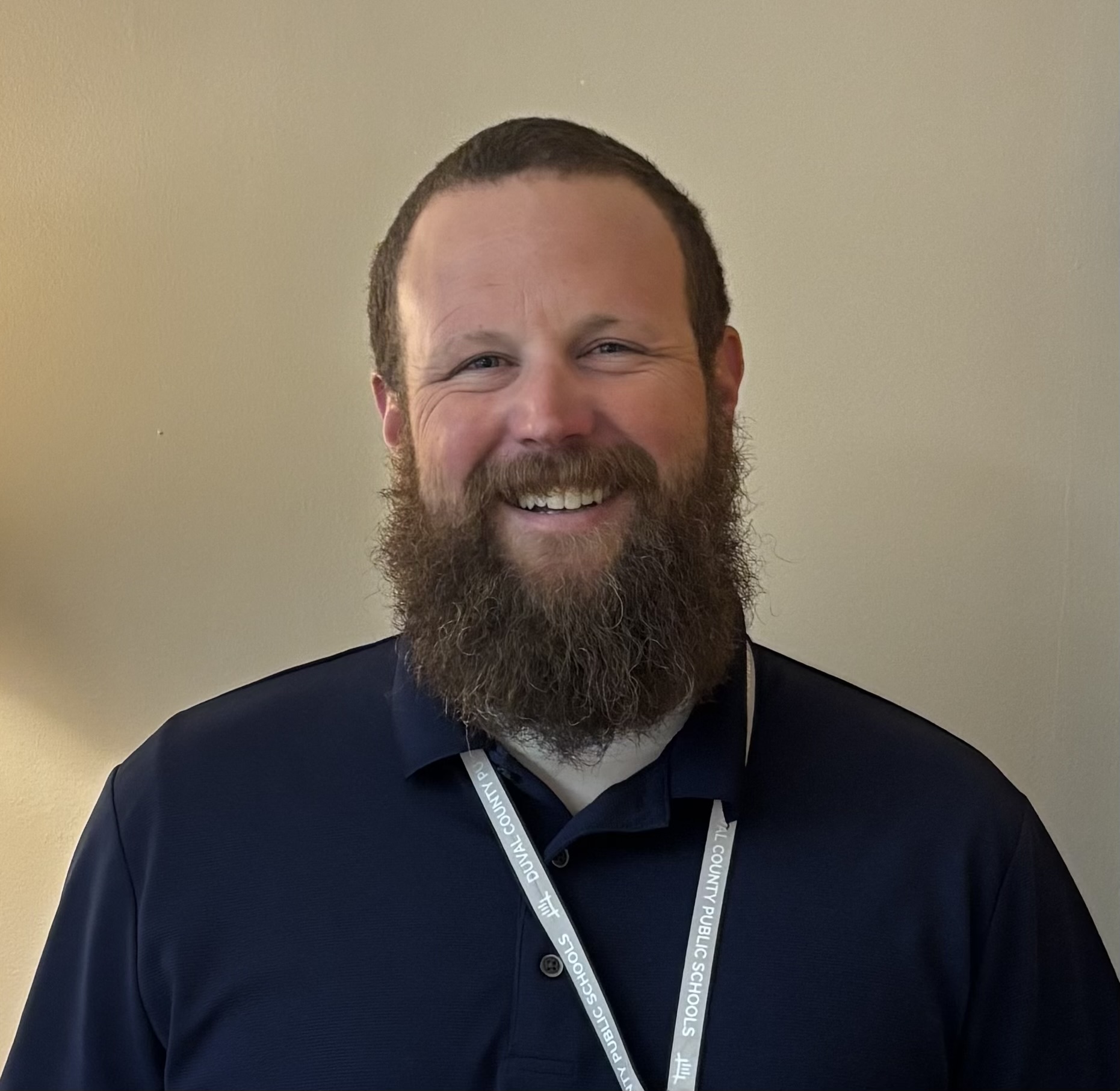 Man with a beard wearing a navy polo and a white lanyard. He smiles and stands in front of a light beige wall.