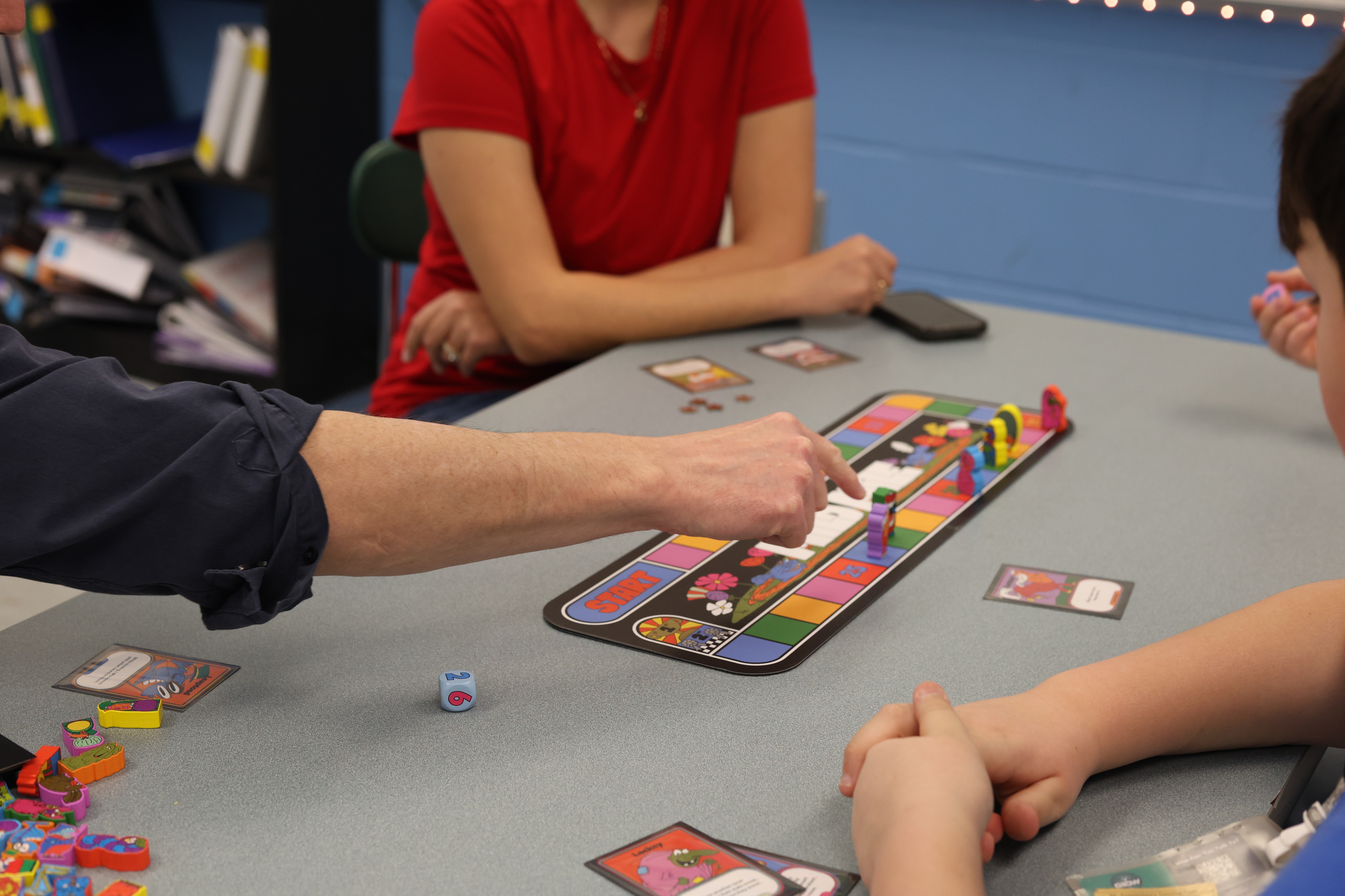 People sitting around a table play a game with colorful cards and dice on a blue wall.