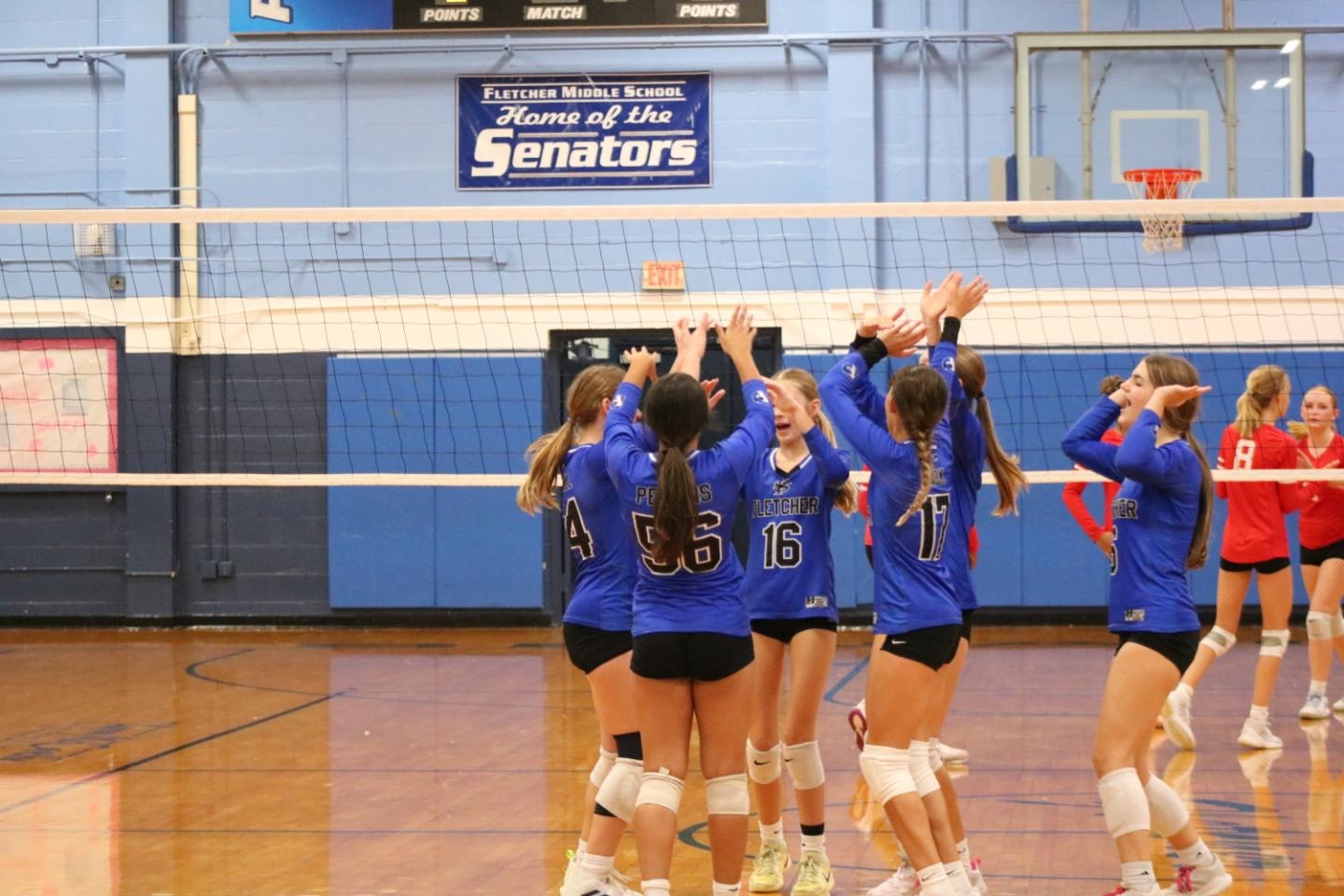 A volleyball team in blue uniforms celebrates together on a court, with a banner reading "Senators" in the background.