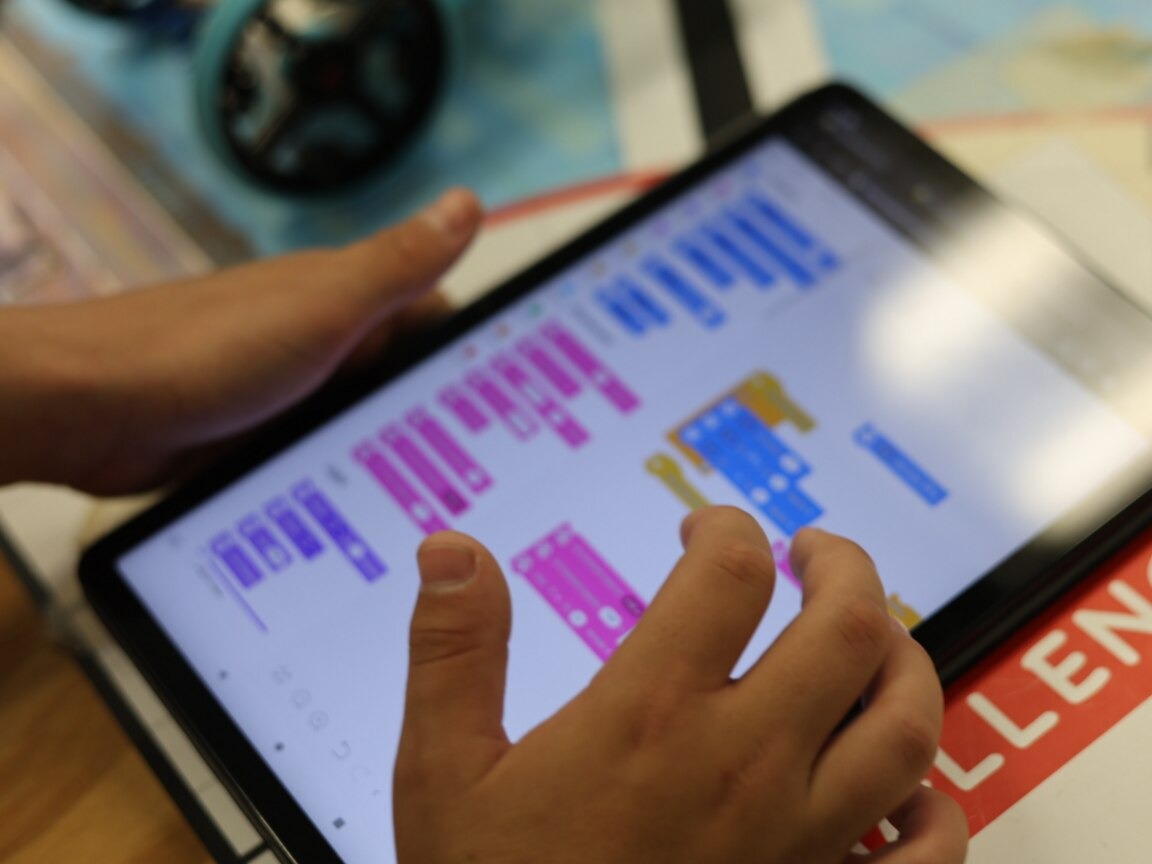 Hands touching a tablet screen displaying a colorful block-based programming interface. A blurred object in the background.