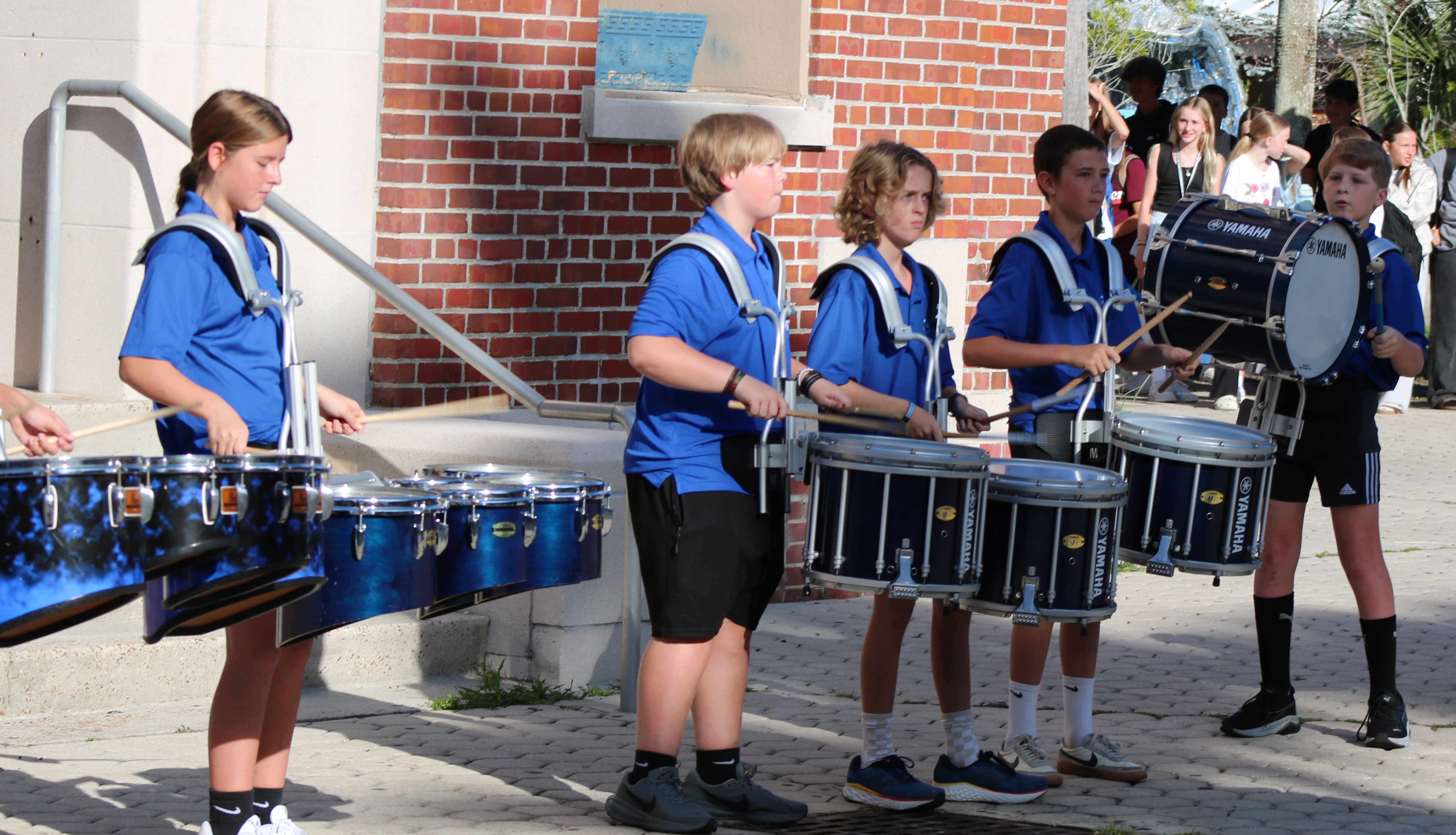 Five children in blue uniforms and black shorts play drums outside a brick building.