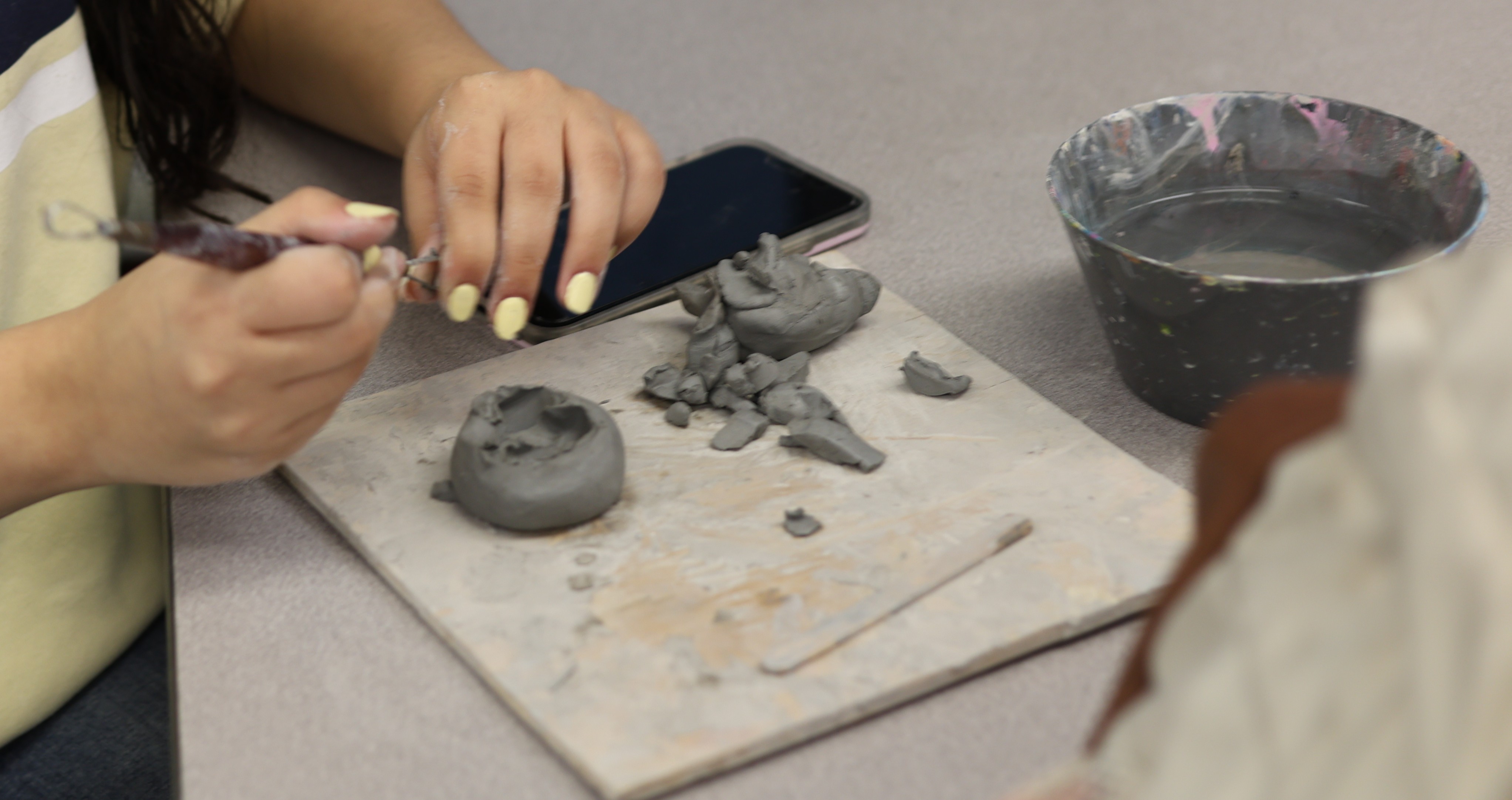 A person is working with clay on a table. Tools and a bowl are nearby.