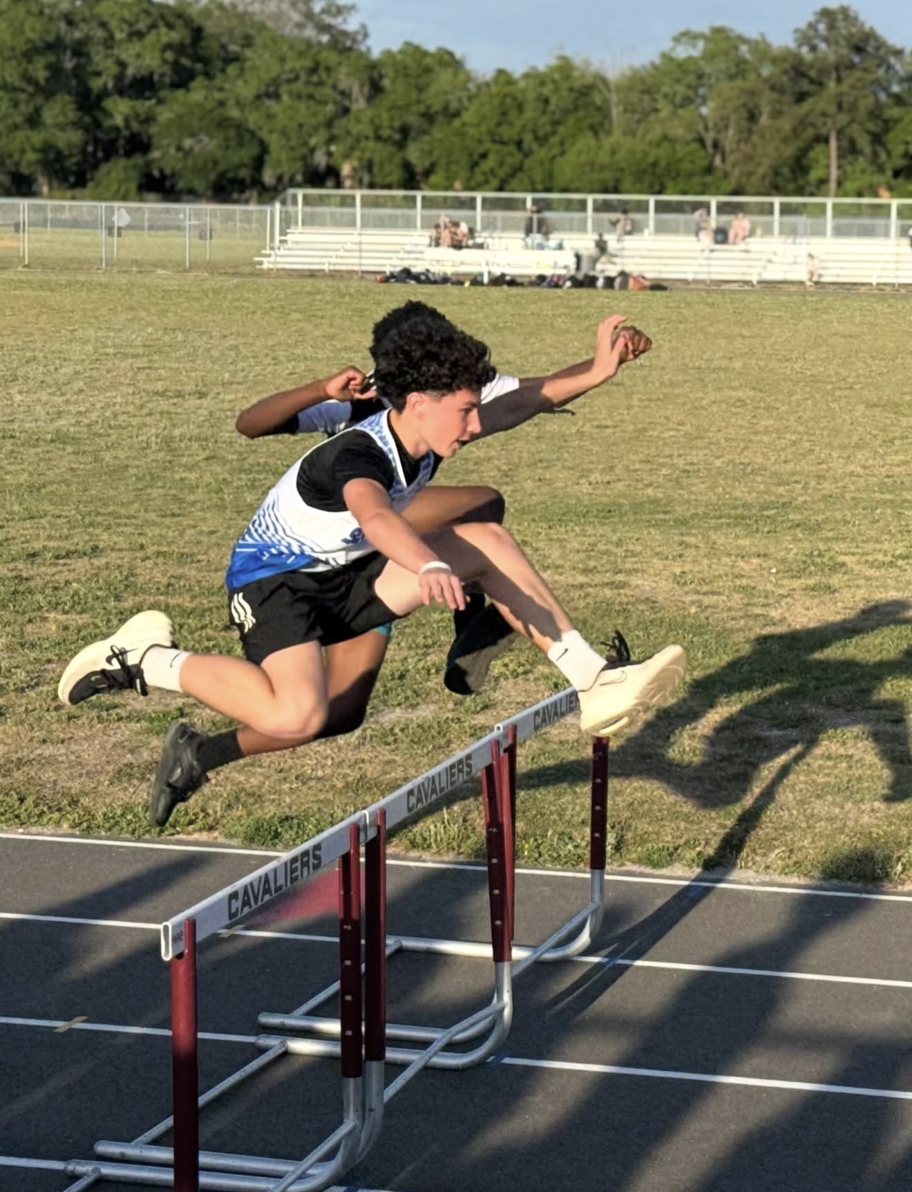 Two athletes jumping hurdles on a track, one in mid-air, with green grass and trees in background.